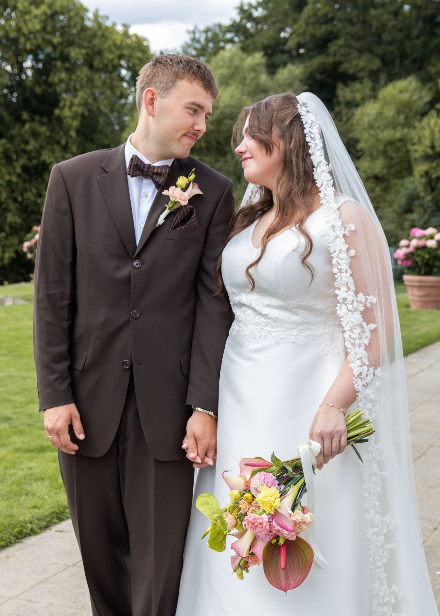 Groom in a brown suit and bow tie gazes at his smiling bride in a lace veil as they hold hands in a leafy garden.