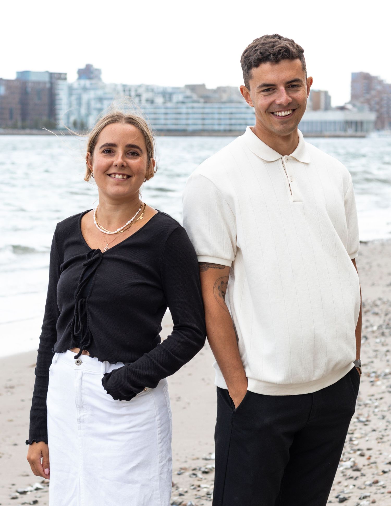 A smiling man in a cream polo and a woman in a black top and pearl necklace pose together by the sea with buildings behind.