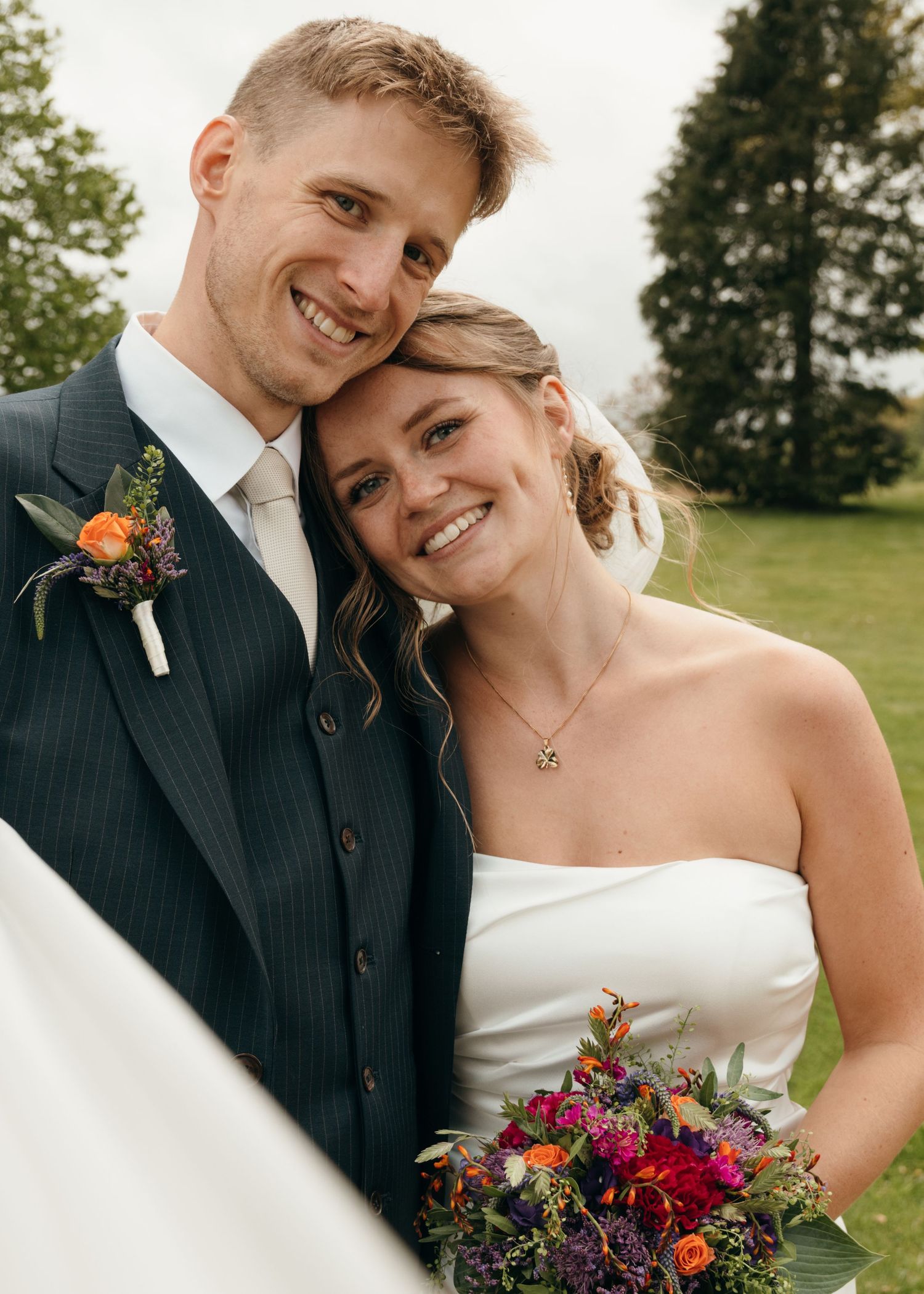 Smiling groom with an orange boutonniere rests his head against his bride, who holds a colorful wedding bouquet outdoors.