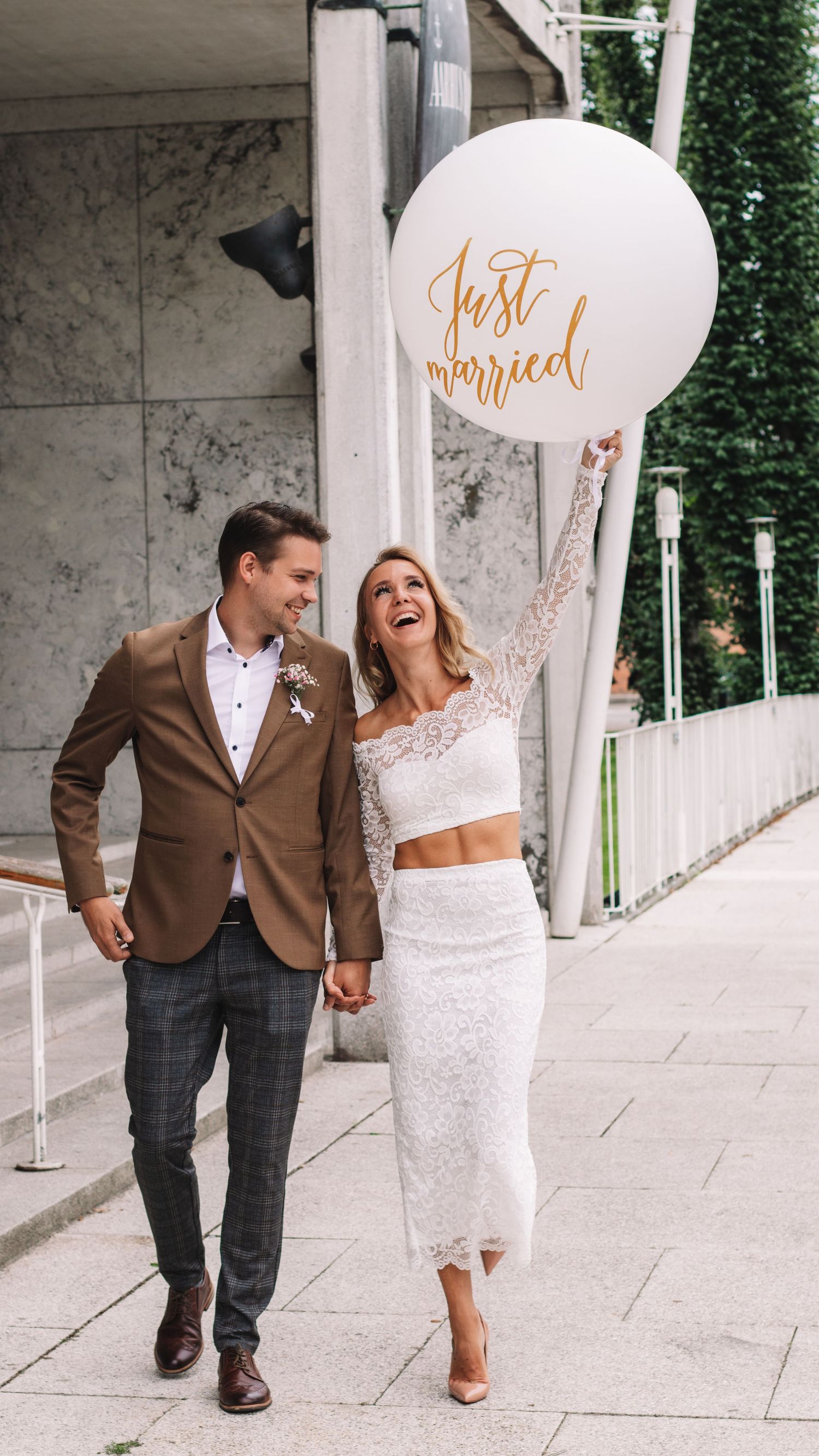 Smiling newlywed couple holding hands and walking outdoors while the bride holds a large white Just married balloon.