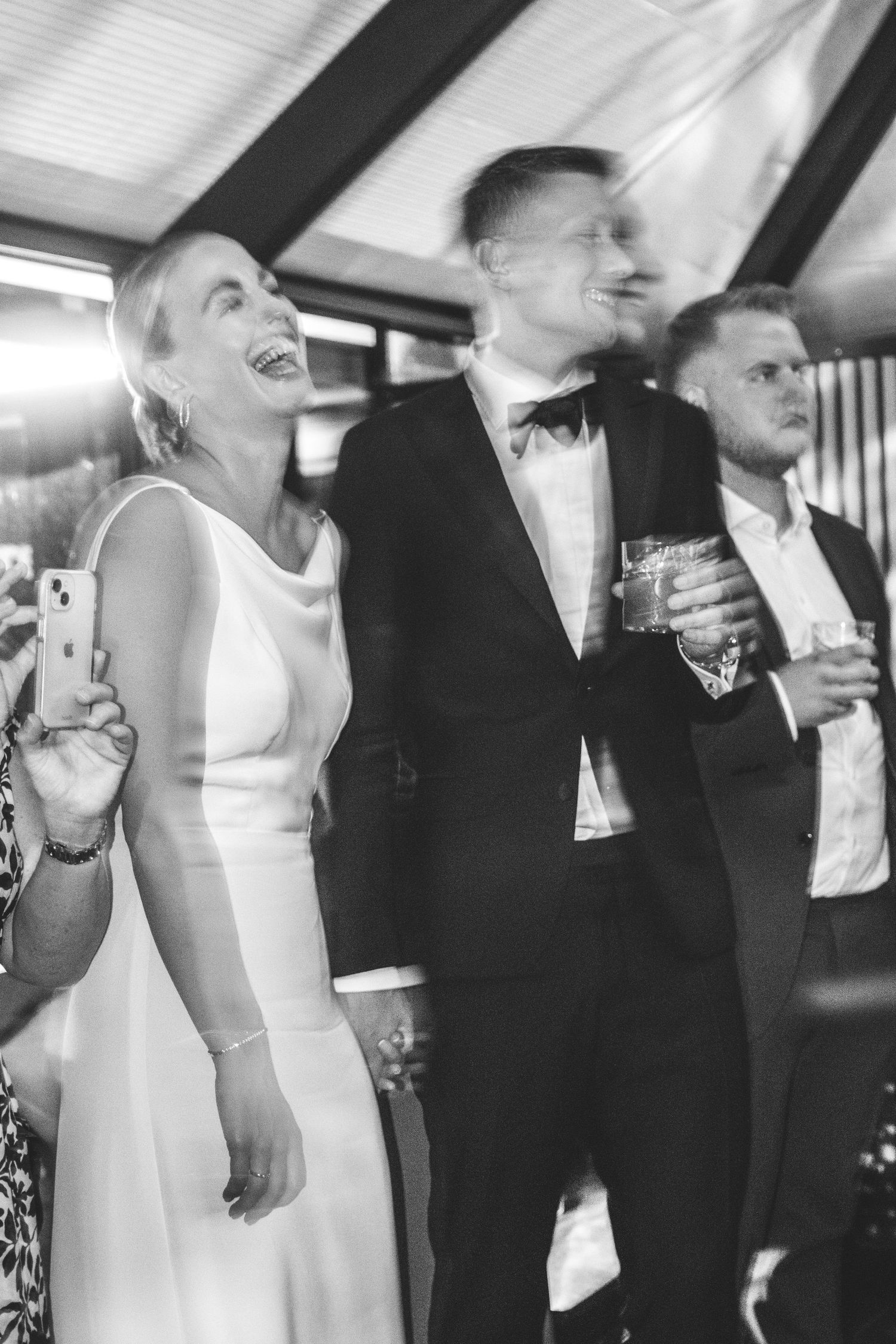 Black and white photo of a bride laughing joyfully beside a groom in a bow tie holding a drink at a wedding reception.