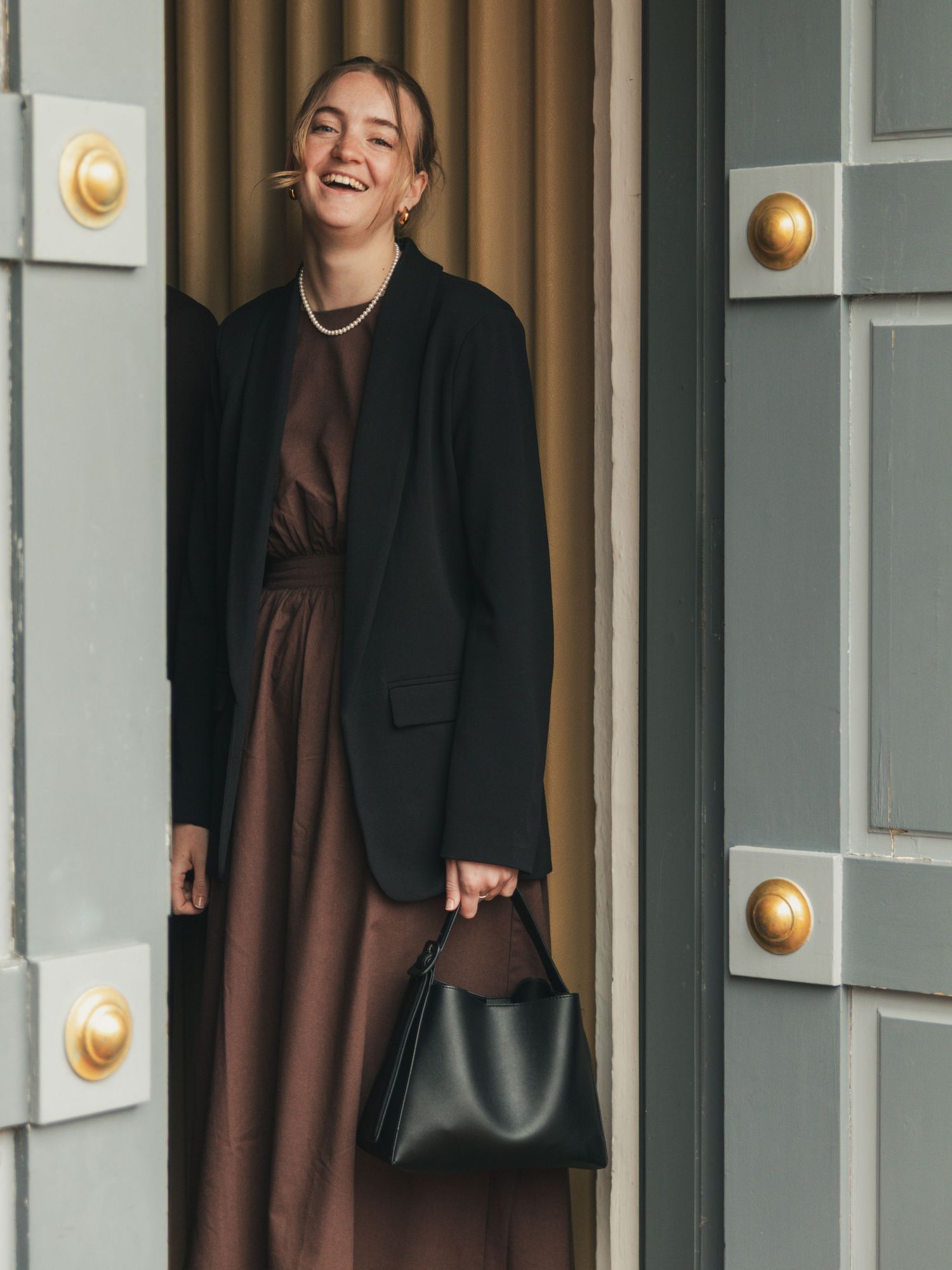 Laughing woman in a black blazer and brown dress with a pearl necklace stands in a doorway holding a black leather handbag.