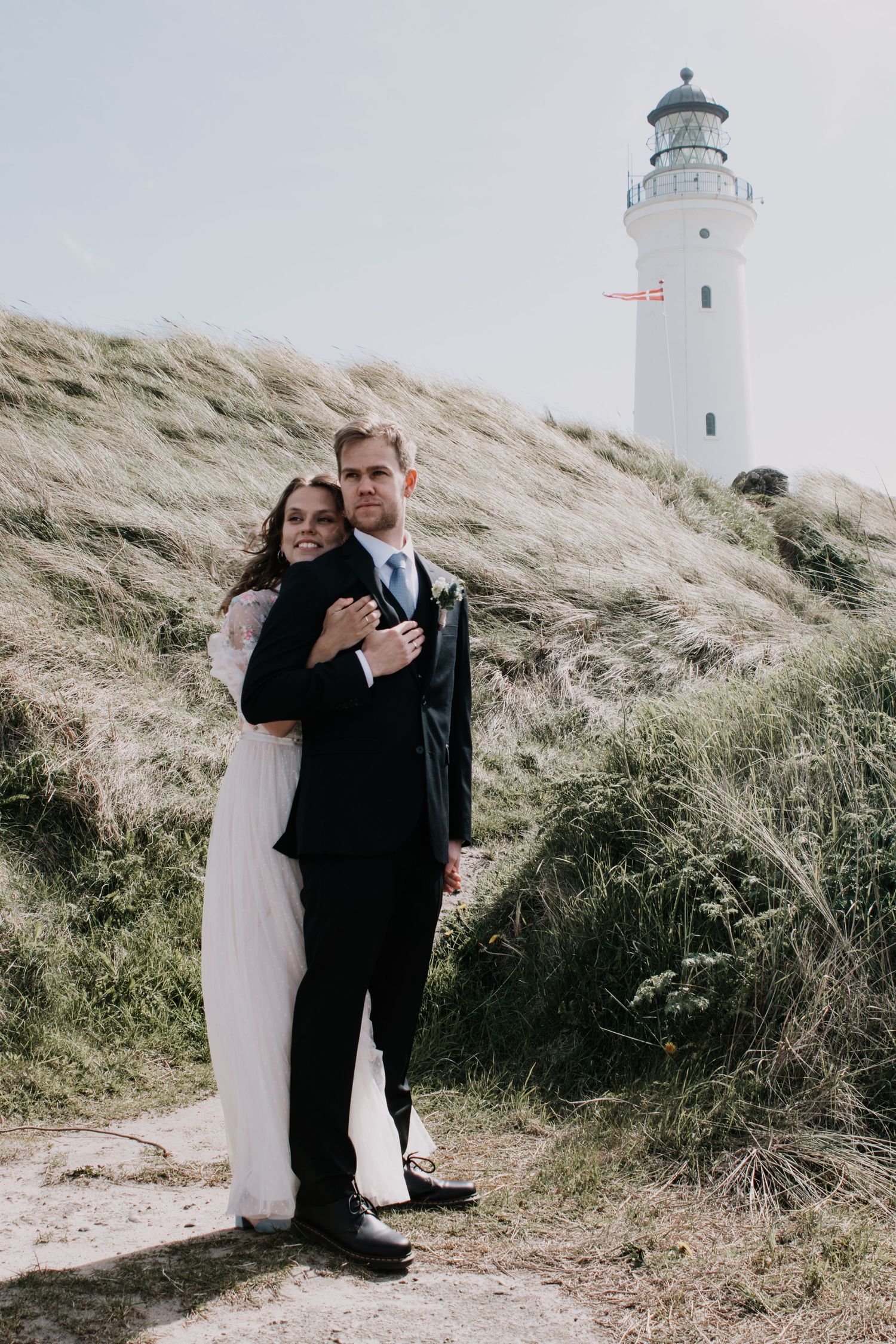 Bride hugs her groom from behind in front of tall dune grass and a white lighthouse flying a small Danish flag.