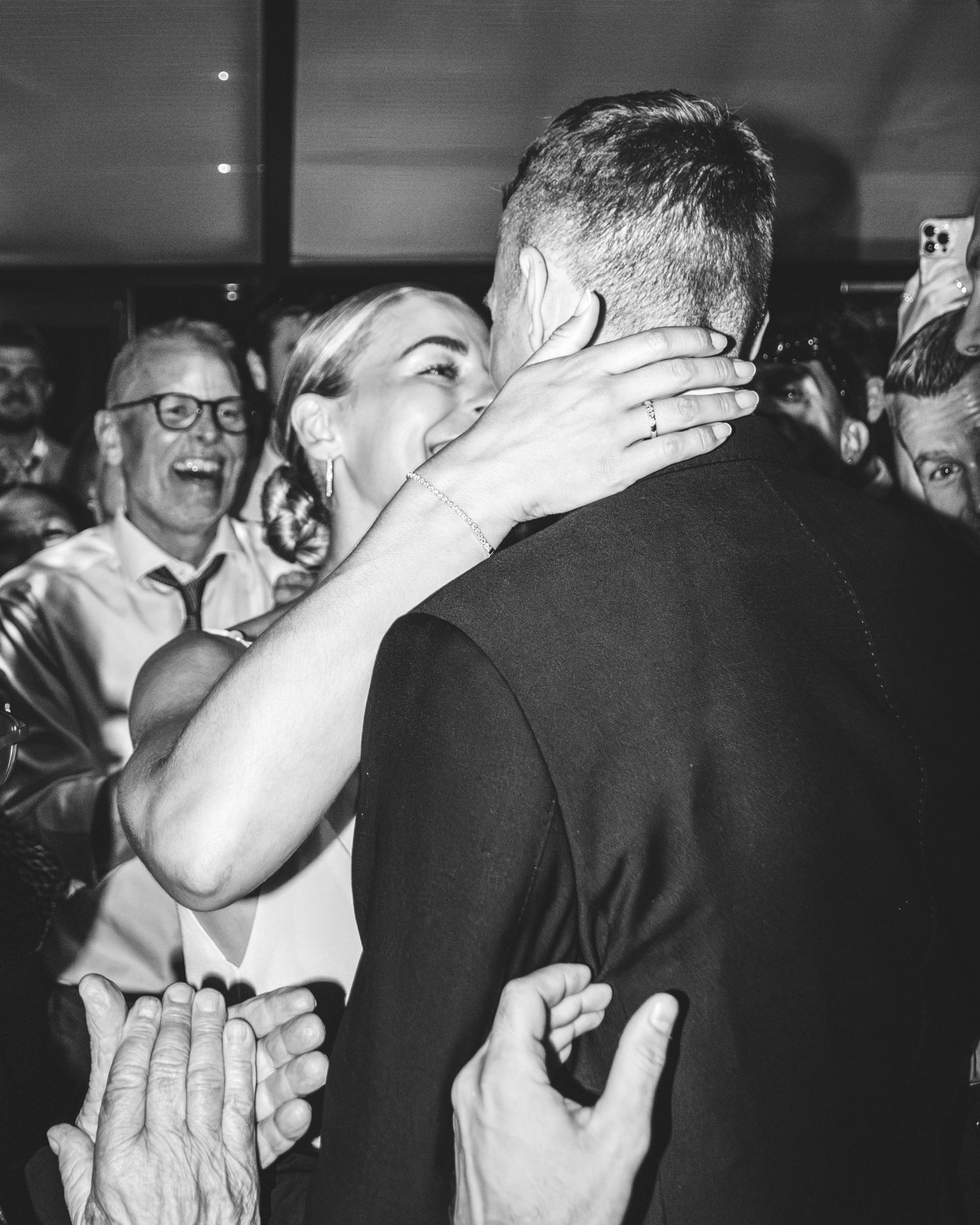Black and white photo of a bride kissing her groom while a laughing guest in glasses and others celebrate behind them.