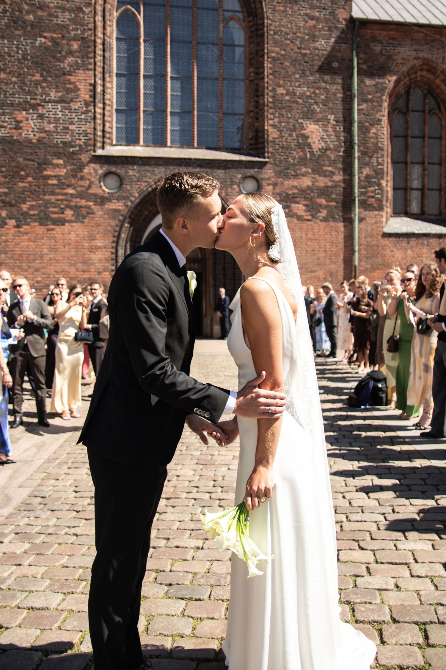 A bride and groom share a wedding kiss on a sunny cobblestone square in front of a brick church as guests gather behind.