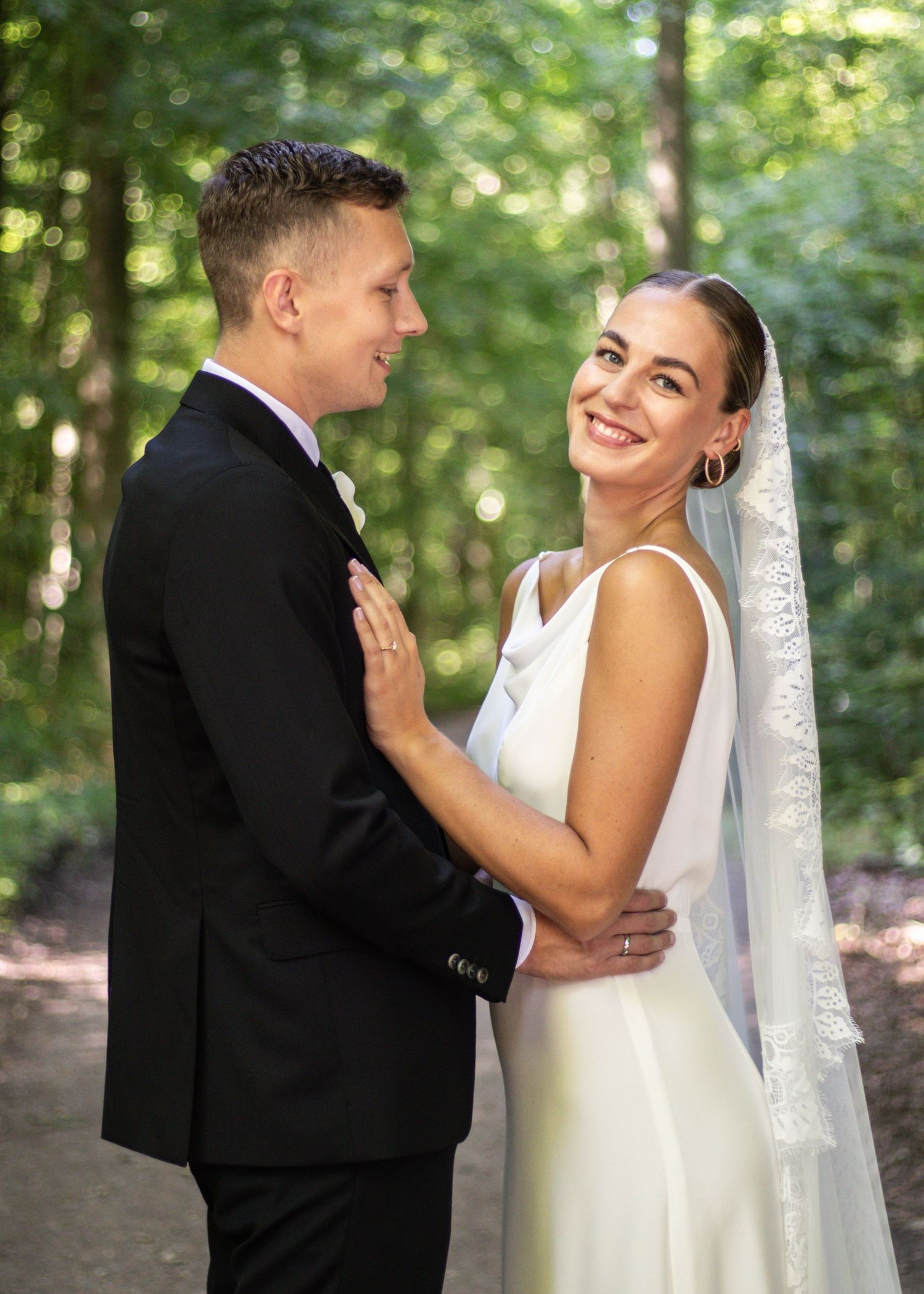 Radiant bride in a white gown and lace veil smiles at the camera while embracing her groom in a black suit on a sunlit forest path.