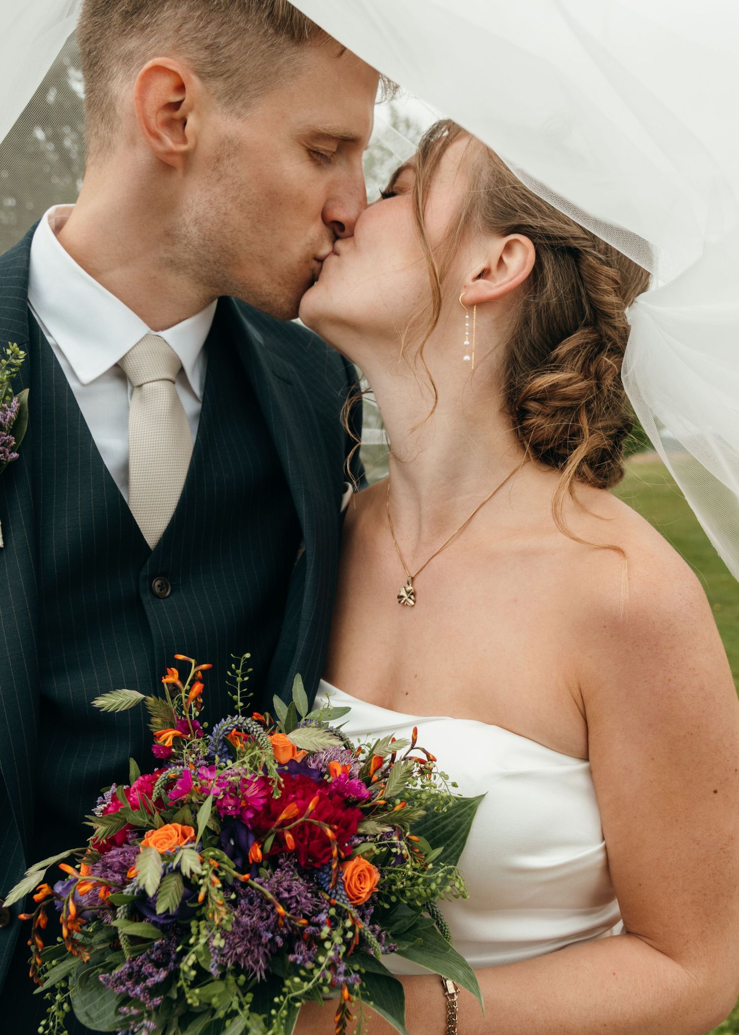 Bride and groom kiss beneath her sheer veil, with a vibrant bouquet of orange, red, and purple flowers in the foreground.