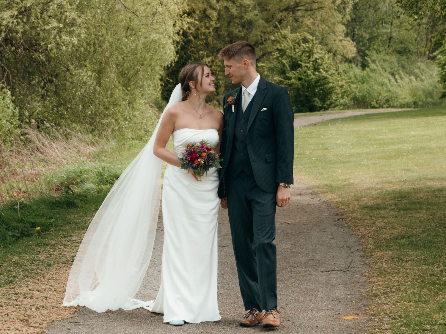 Bride in a strapless white gown and long veil smiles at her groom in a navy suit on a leafy countryside path.