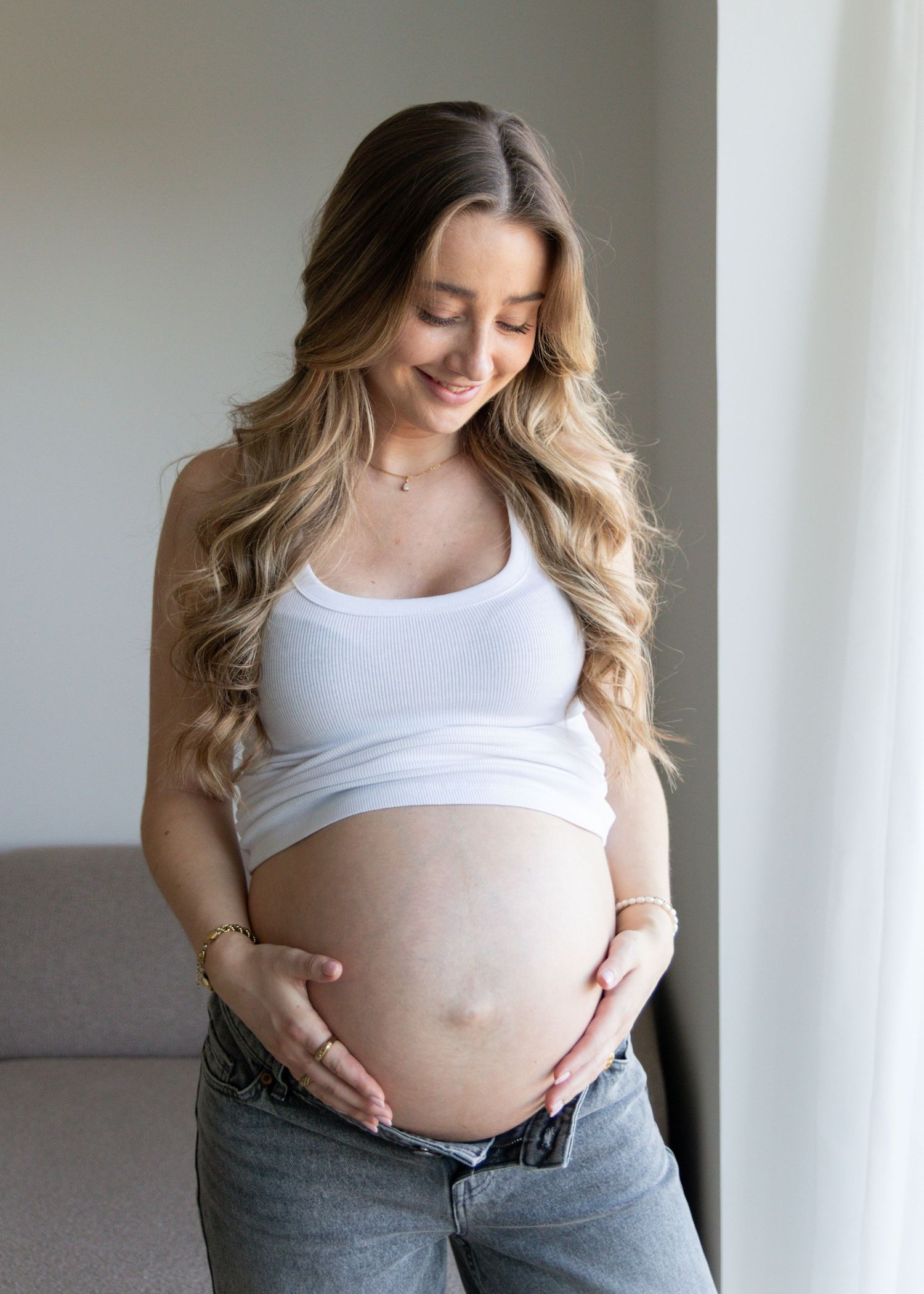 Smiling pregnant woman with long wavy hair in a white crop top gently cradles her bare baby bump in soft natural window light.