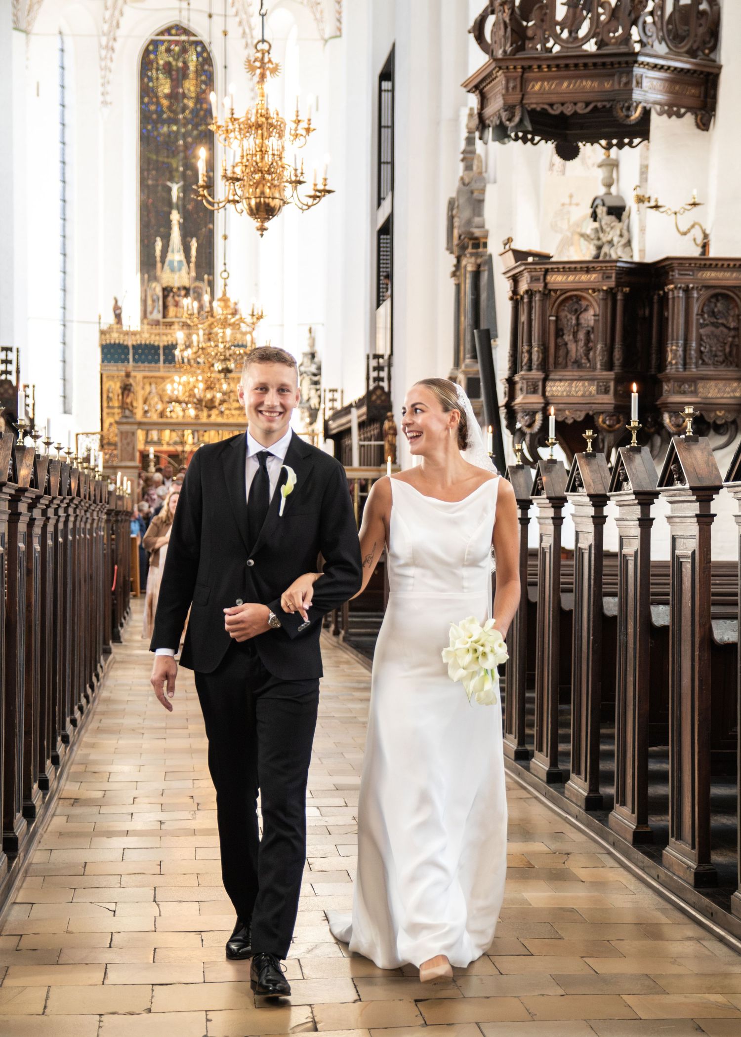 Smiling bride in a white gown holding a bouquet walks arm in arm with the groom down a church aisle beneath gold chandeliers.