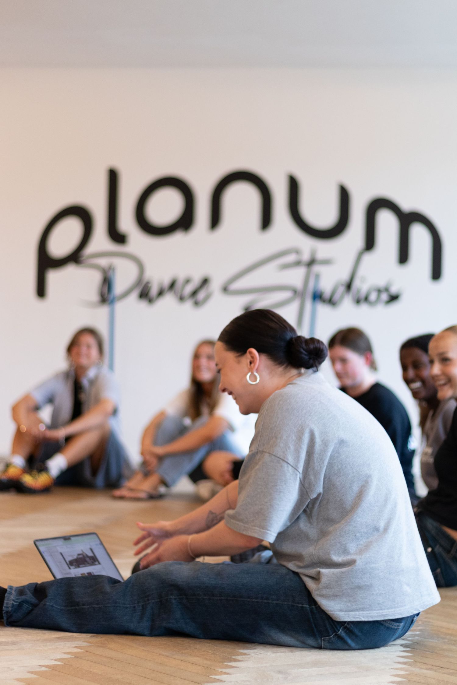 Instructor with a laptop sits on the floor facing a group of students in front of the Planum Dance Studios wall logo.