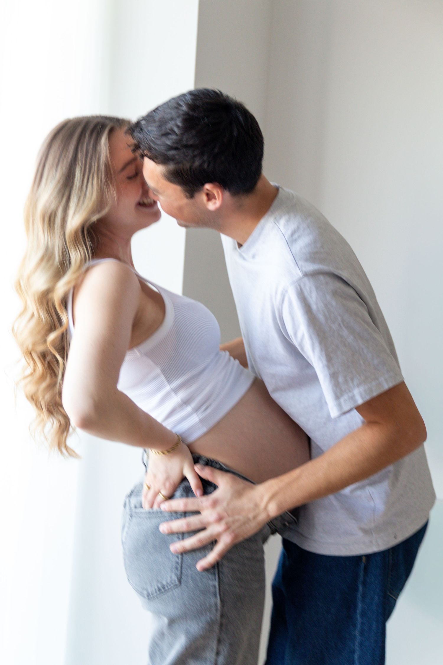A man and a pregnant woman in a white top and jeans share an affectionate moment in a bright, softly lit room.