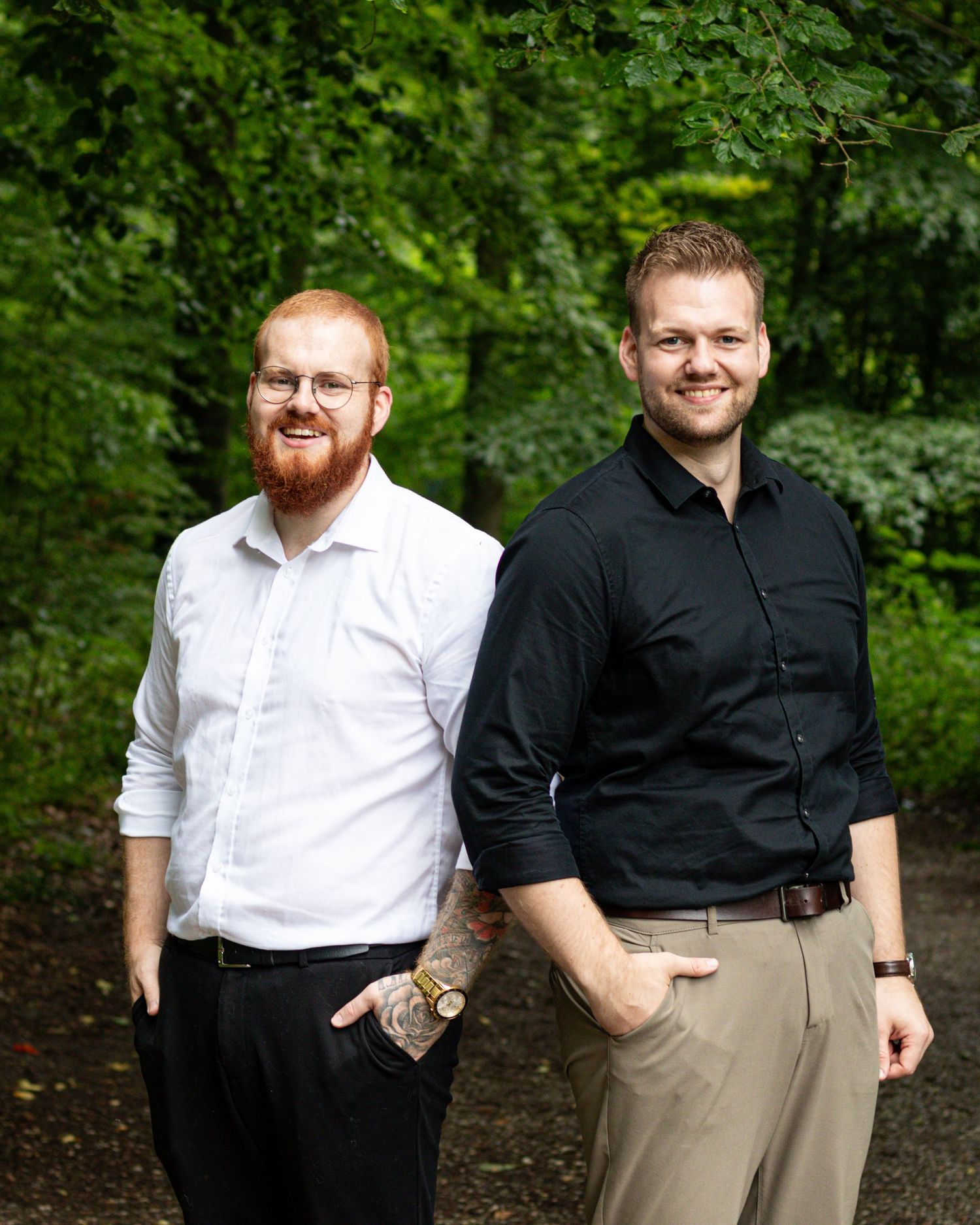 Two smiling men, one with a red beard and glasses in a white shirt, pose together in a lush green forest setting.