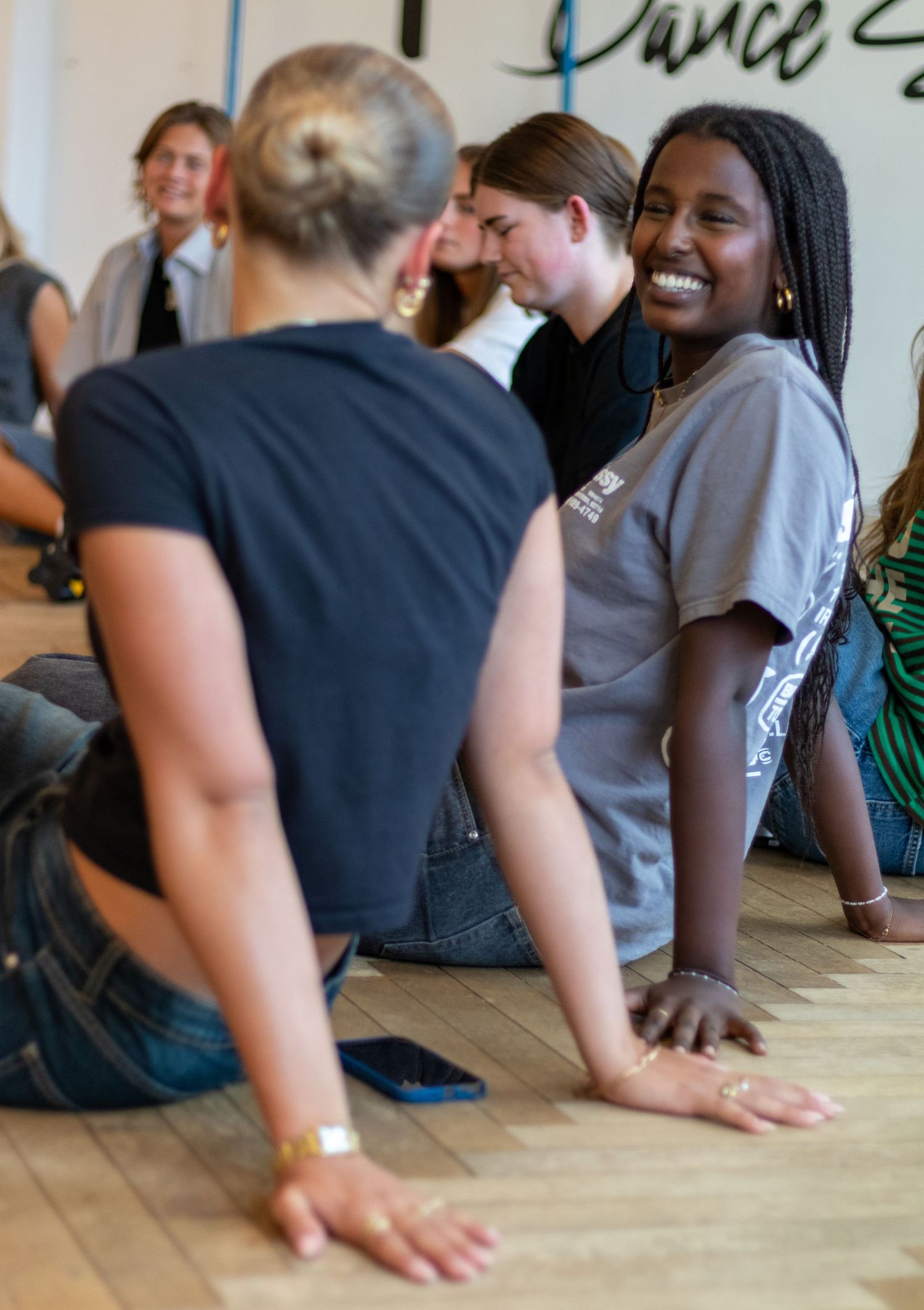 A smiling young woman sits on a wooden floor with other participants during a dance class in a bright studio.
