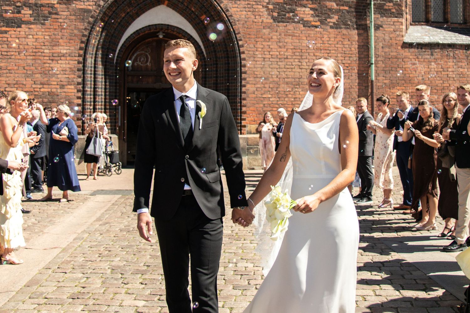 A happy groom in a black suit and a bride holding a bouquet walk hand in hand outside a brick church on a sunny day.