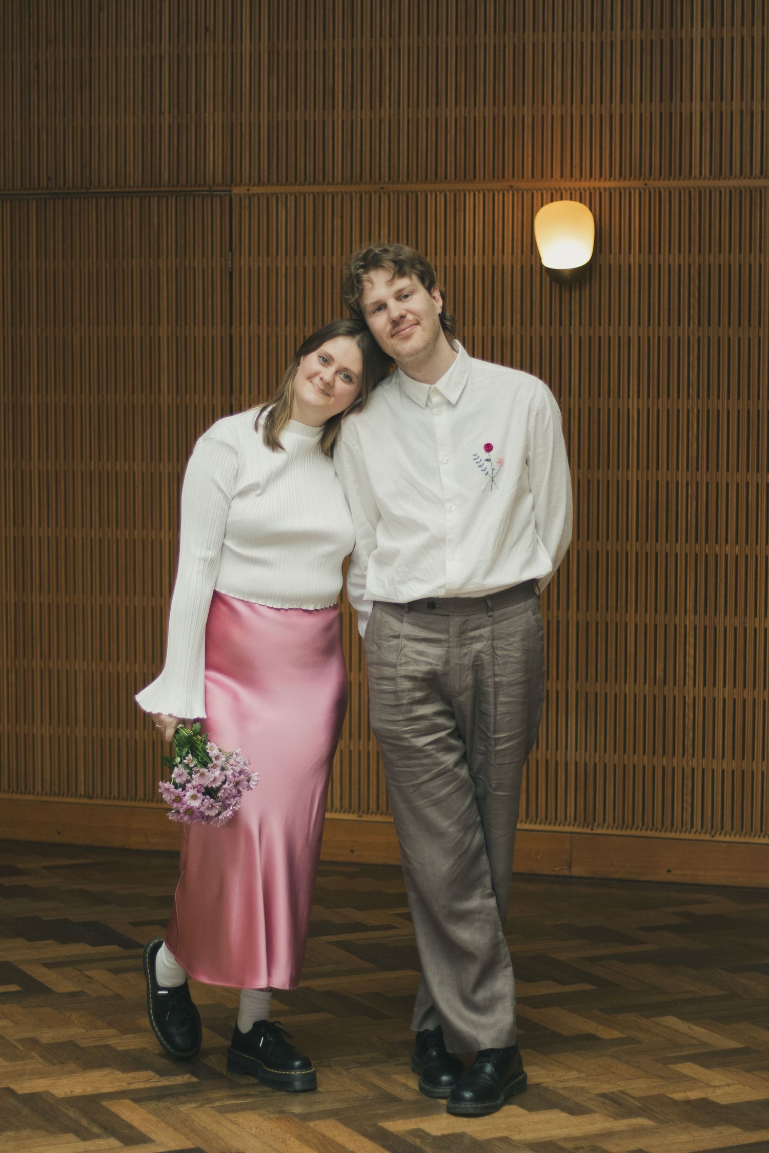 A woman in a pink satin skirt leans her head against her partner's shoulder in a warm wood-paneled room, holding a flower bouquet.