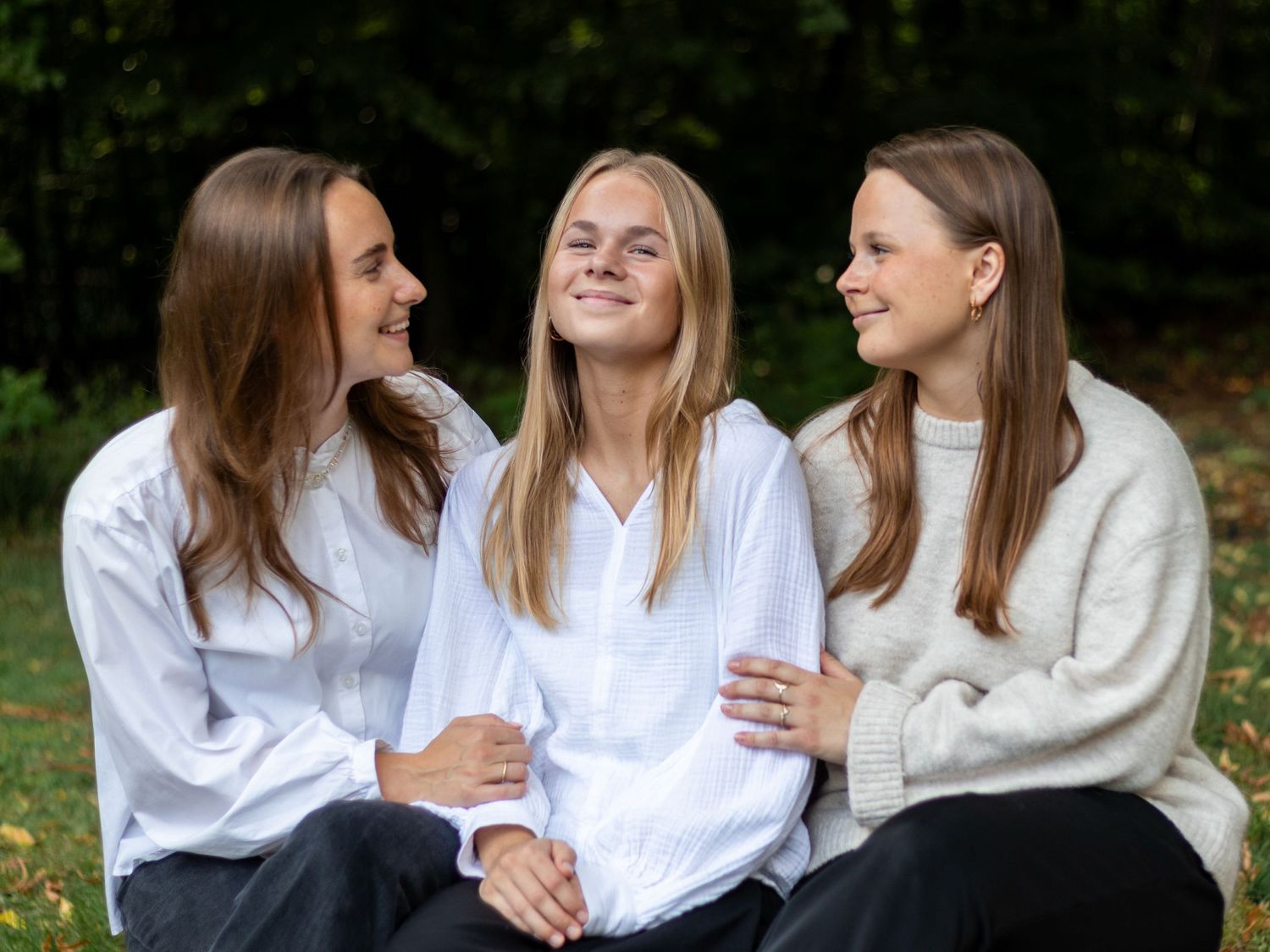 Three young women in white shirts and a cream sweater sit close together on grass, smiling and laughing outdoors.