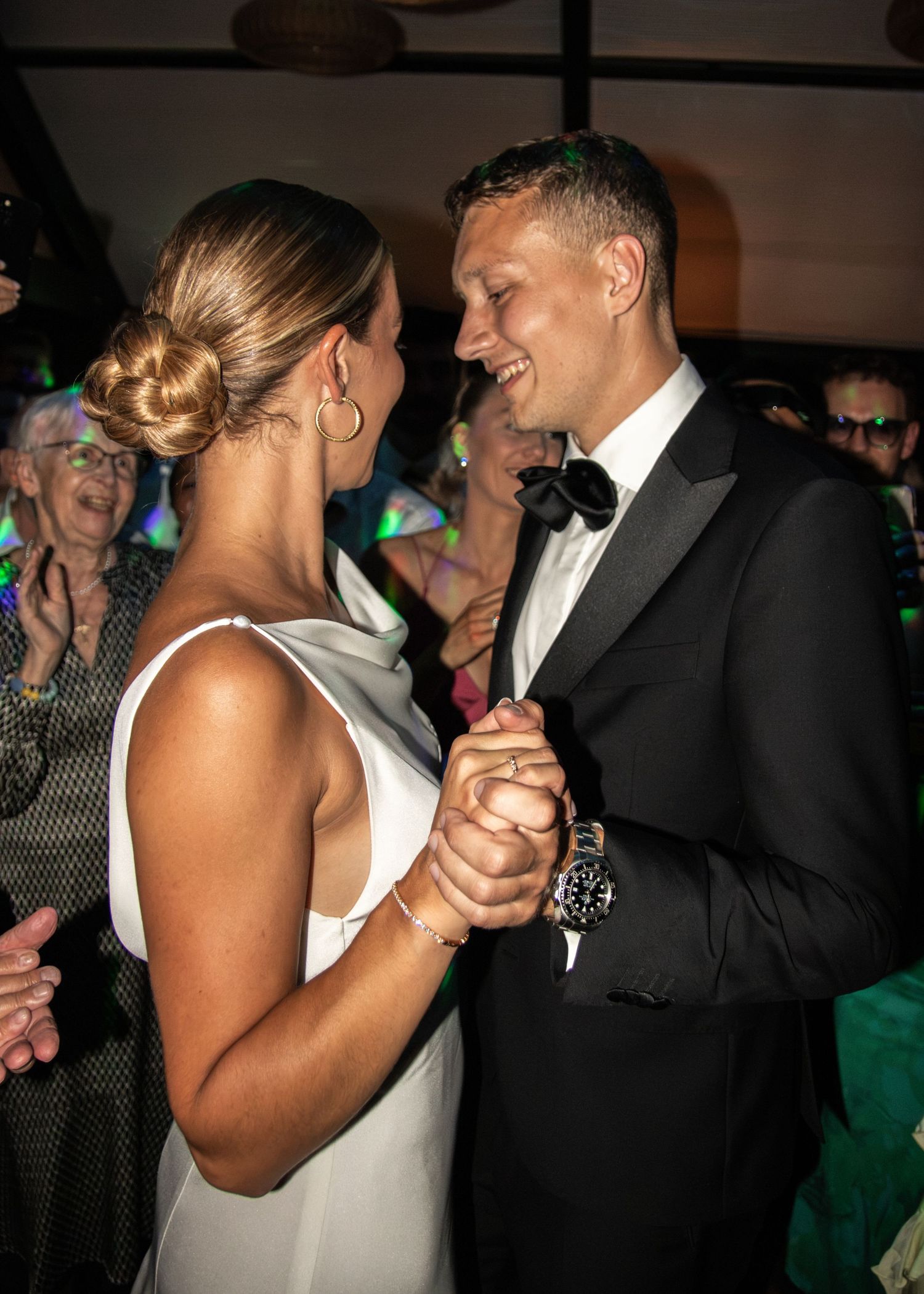 Bride with an elegant updo and groom in a bow tie hold hands and smile at each other as guests applaud at their reception.