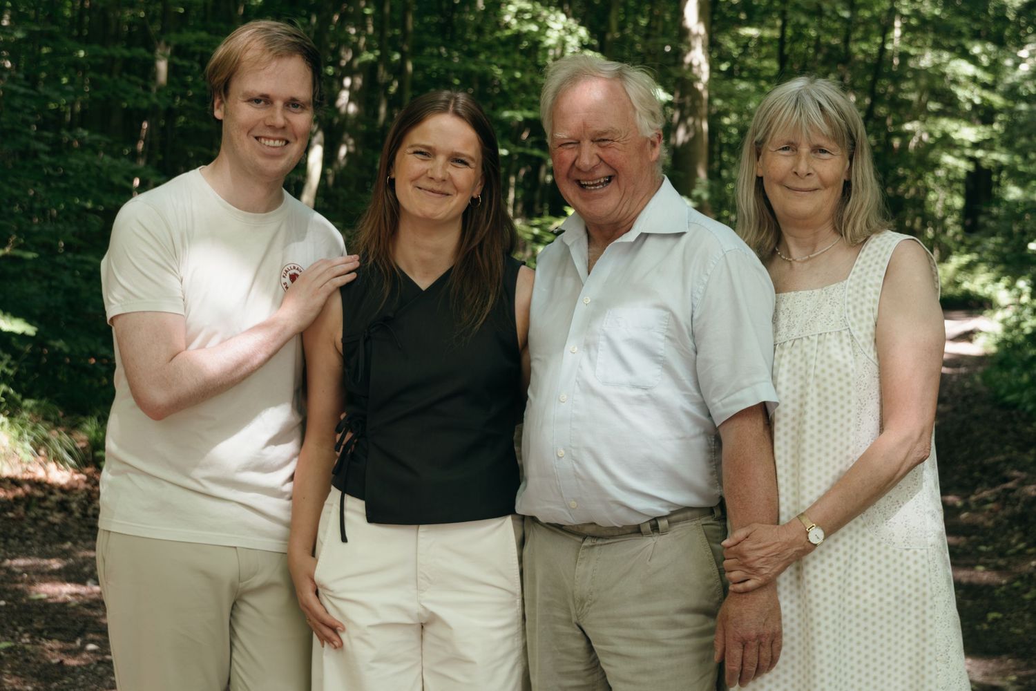 Two men and two women pose together for a happy family portrait on a sunny forest path surrounded by green trees.