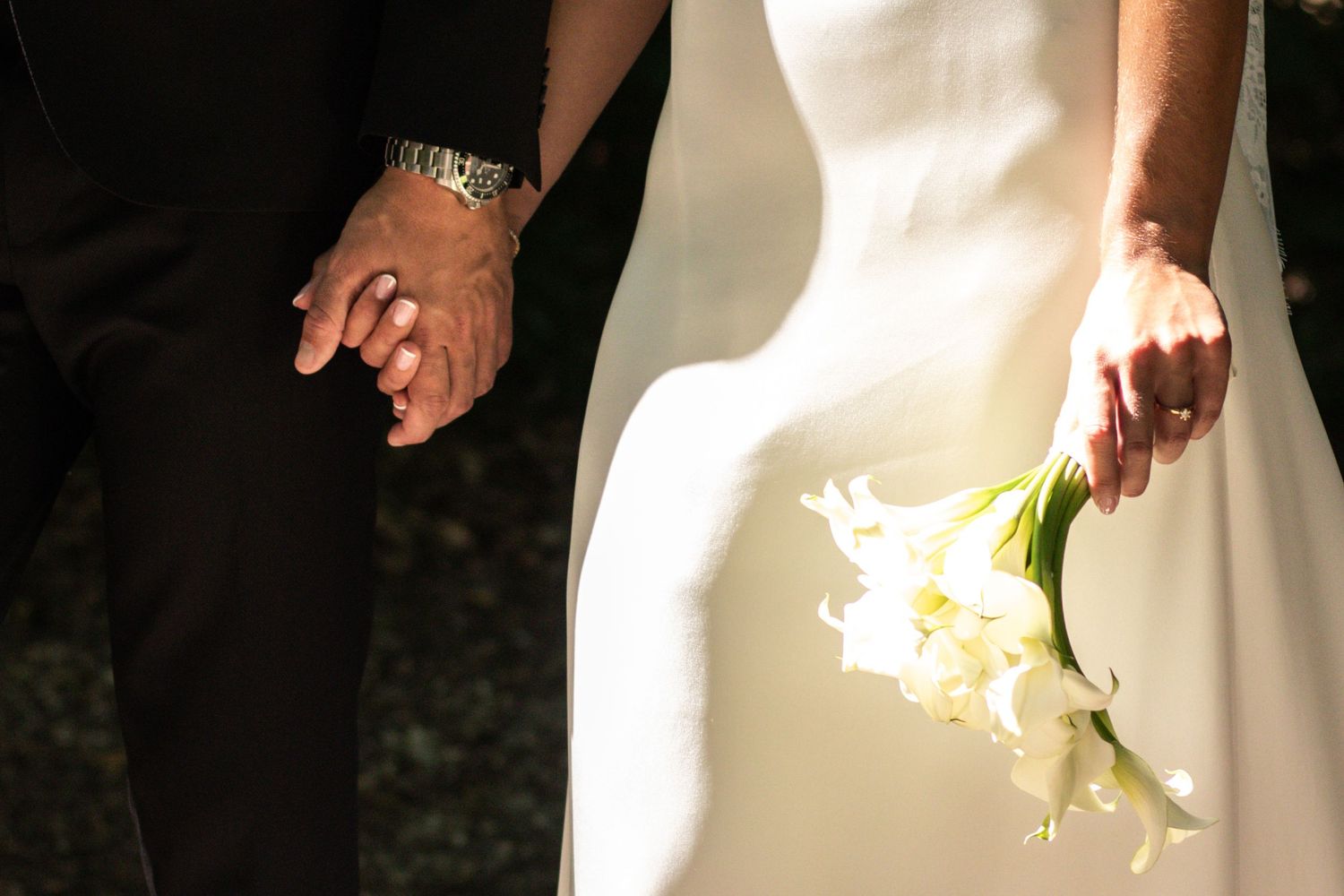 A bride and groom hold hands, wearing wedding rings, as the bride carries a bouquet of white calla lilies.