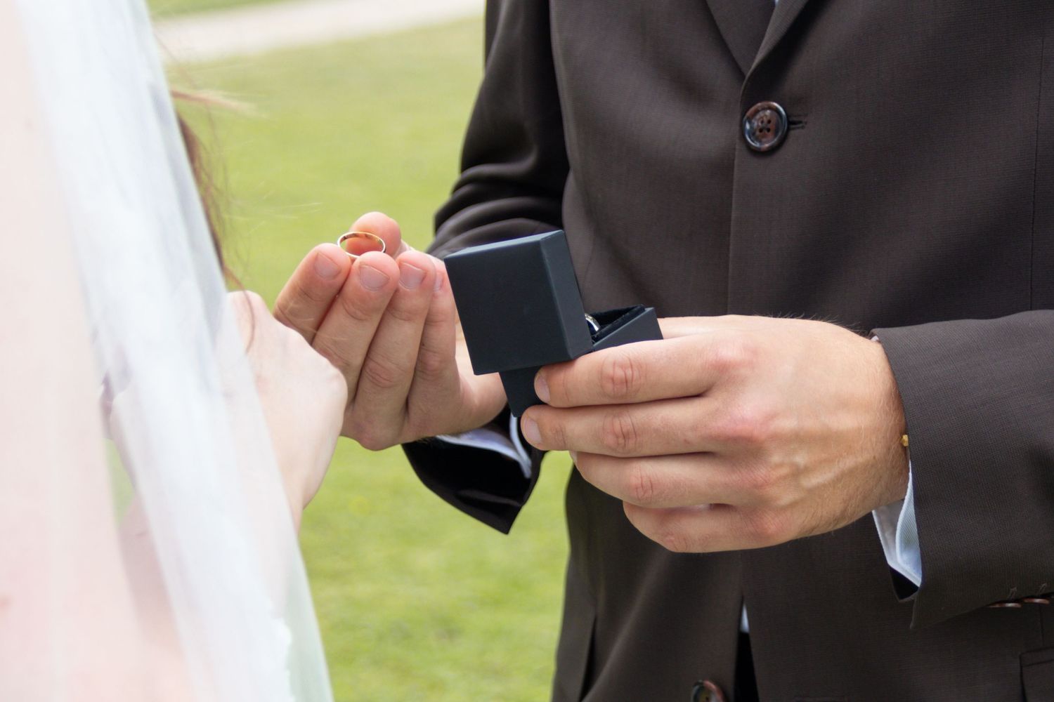 A groom in a dark suit holds an open black ring box toward the bride's hand during an outdoor wedding ceremony on a green lawn.