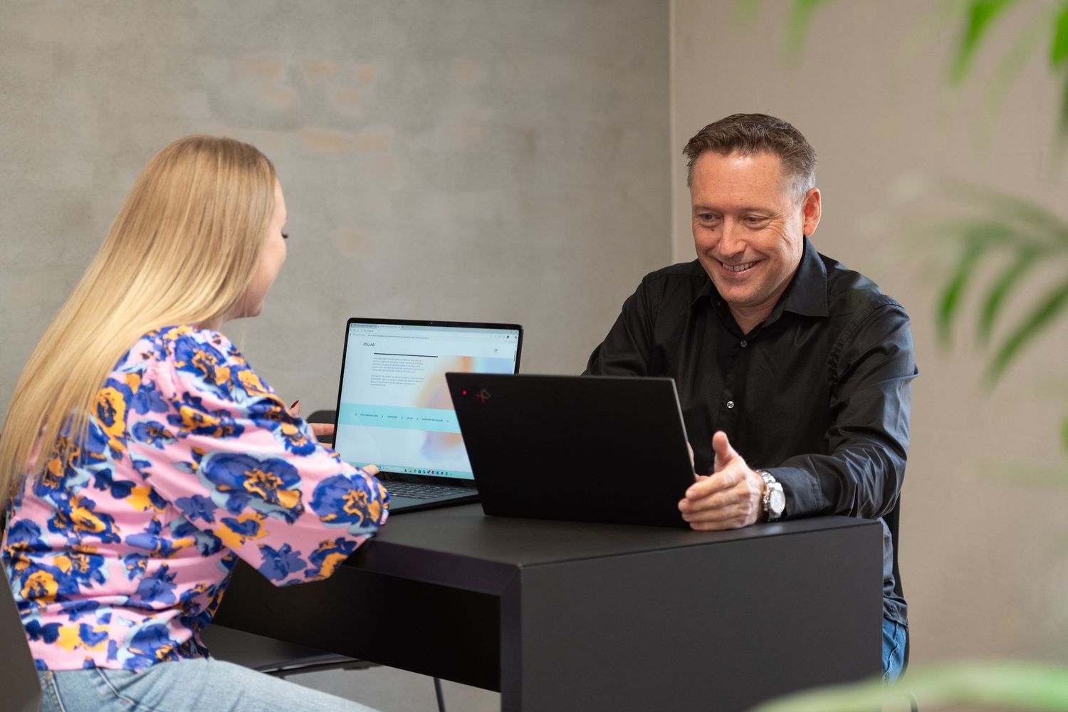 A smiling man in a black shirt talks with a woman in a colorful floral top across a dark desk with laptops.