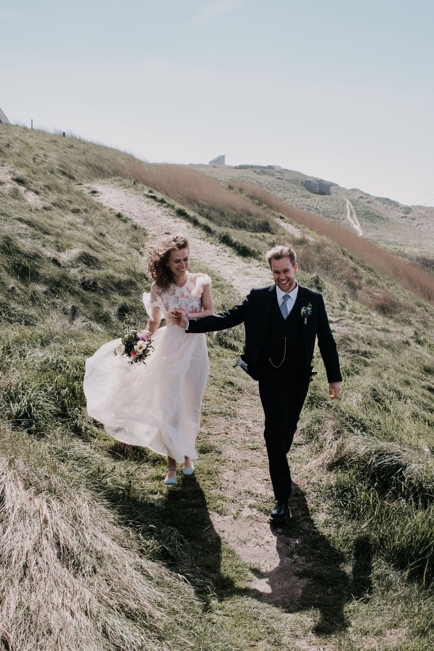 Smiling bride with a bouquet and groom in a dark suit walk hand in hand along a grassy hillside path on a sunny day.