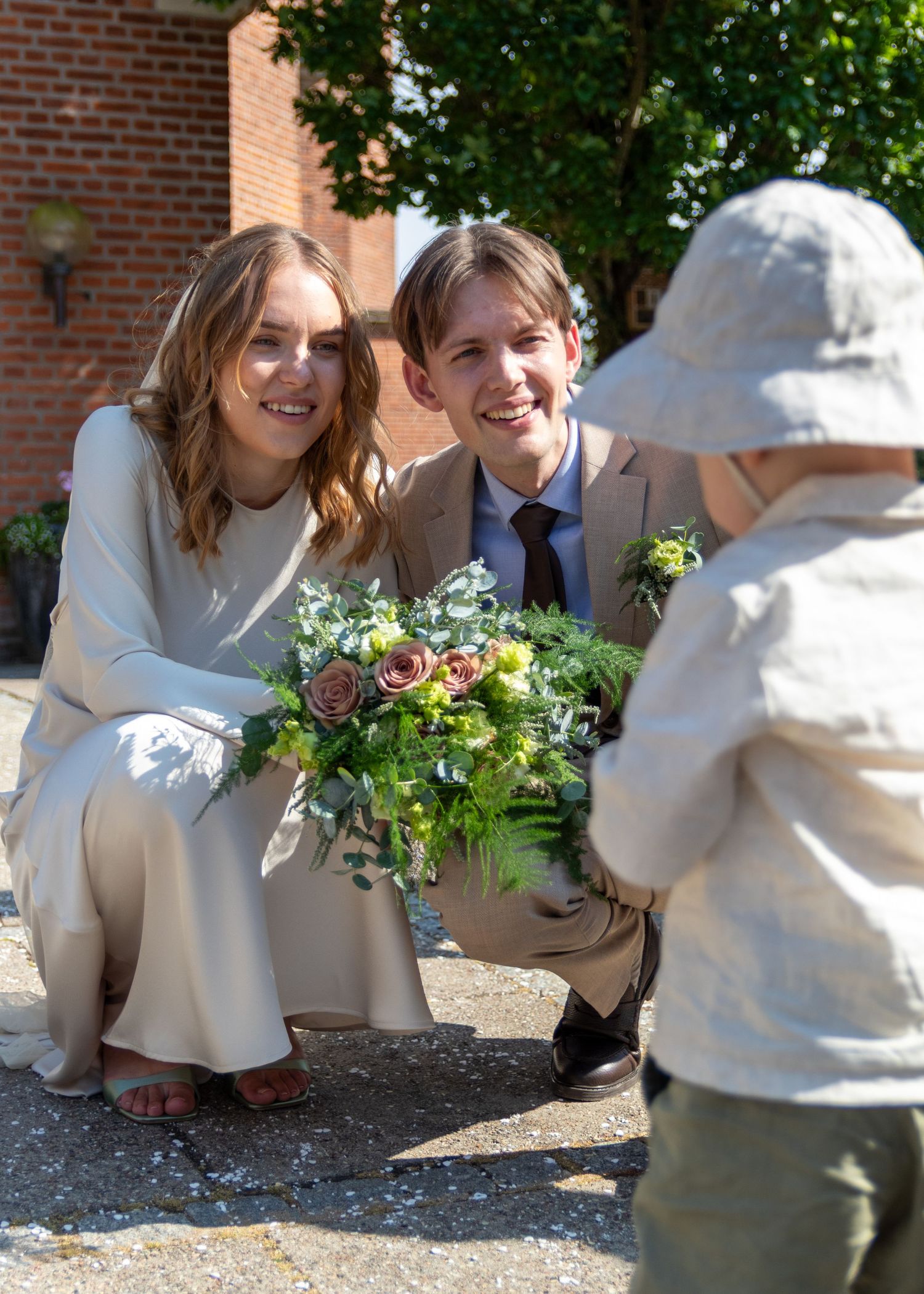 Bride and groom crouch down smiling at their toddler in a linen sun hat, holding a wedding bouquet outdoors.