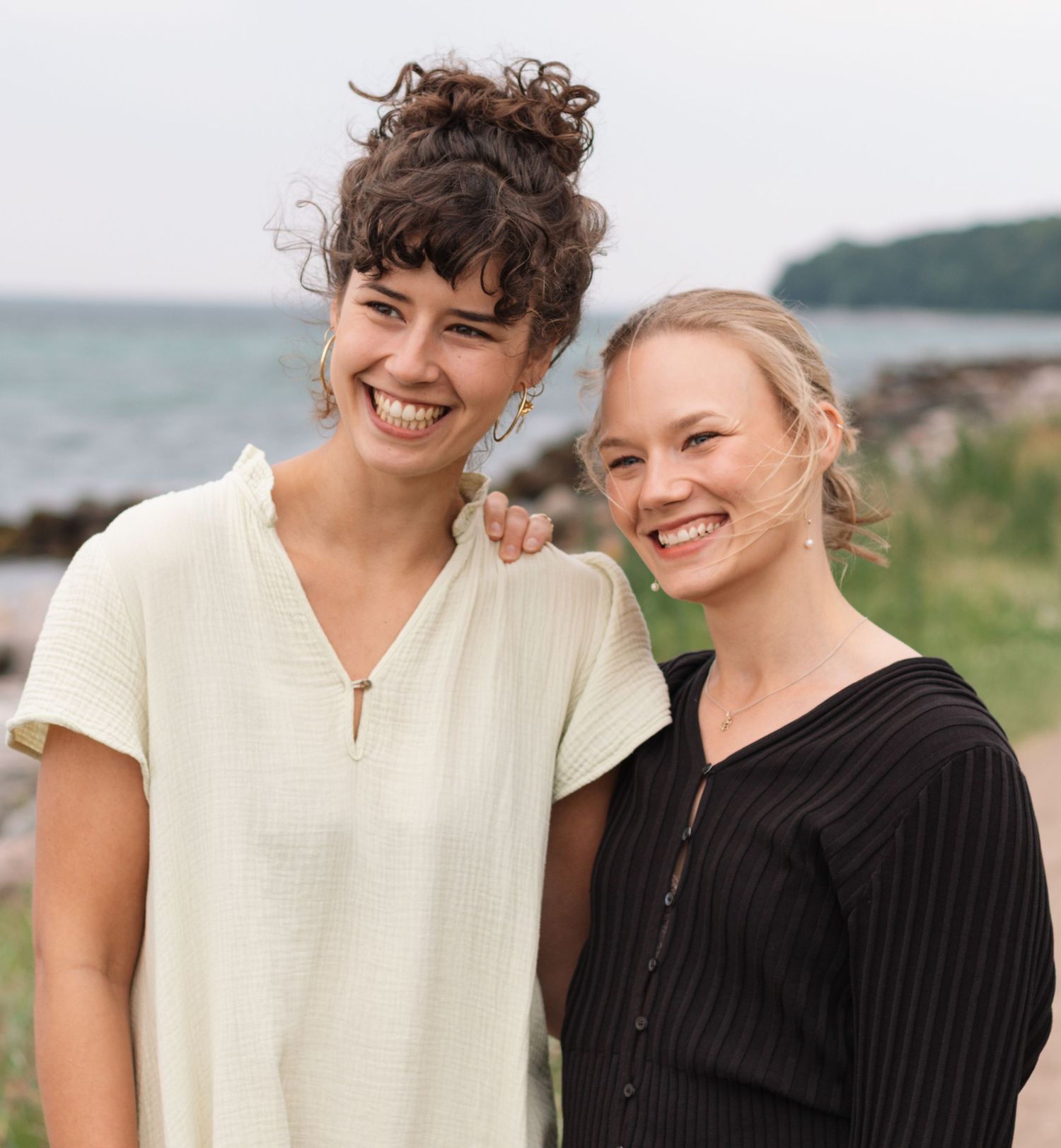 Two women laugh together in a close portrait outdoors, with a rocky shoreline and sea softly blurred behind them.