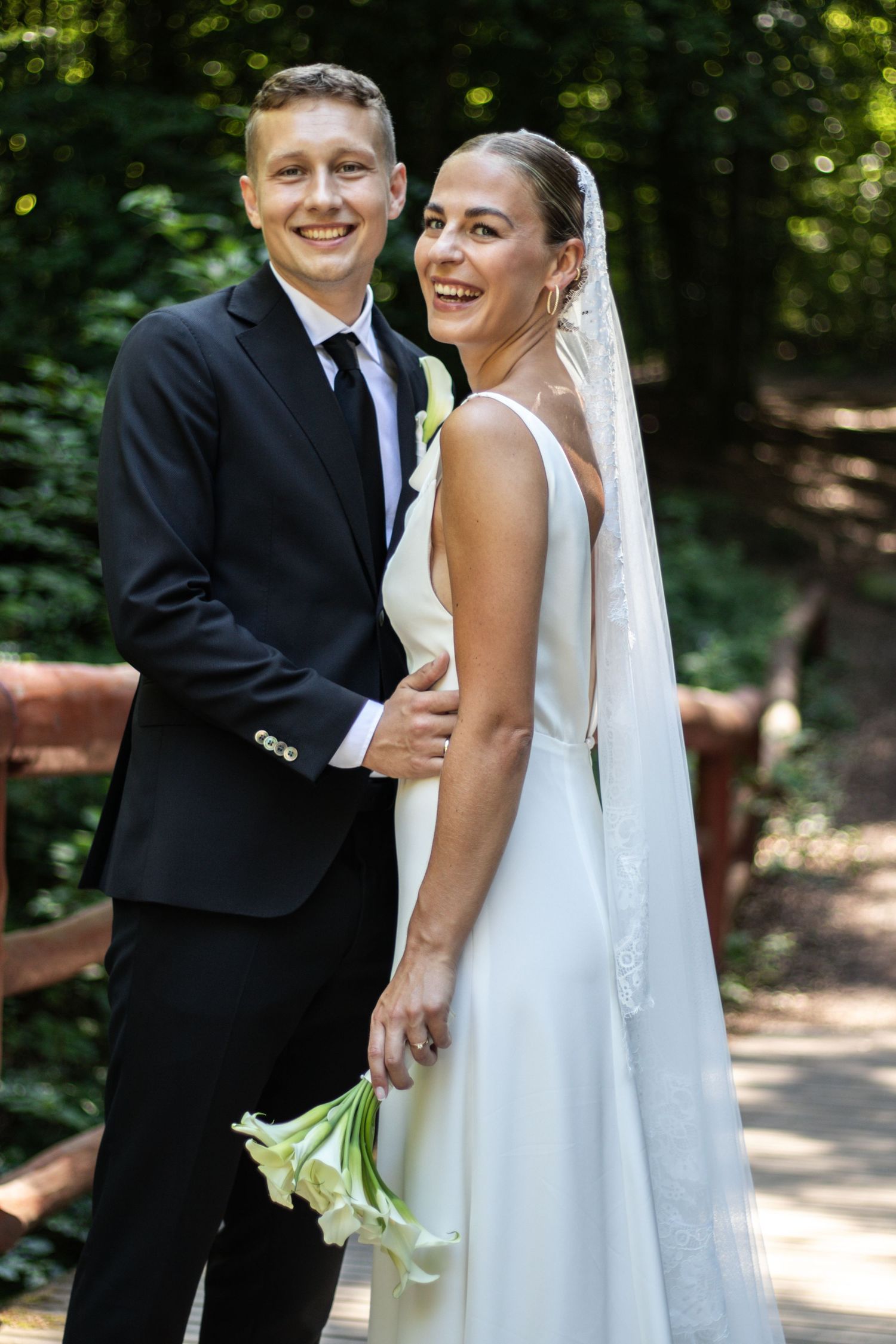 Laughing bride in a white wedding gown and long veil holds a white calla lily bouquet beside her smiling groom on a forest trail.