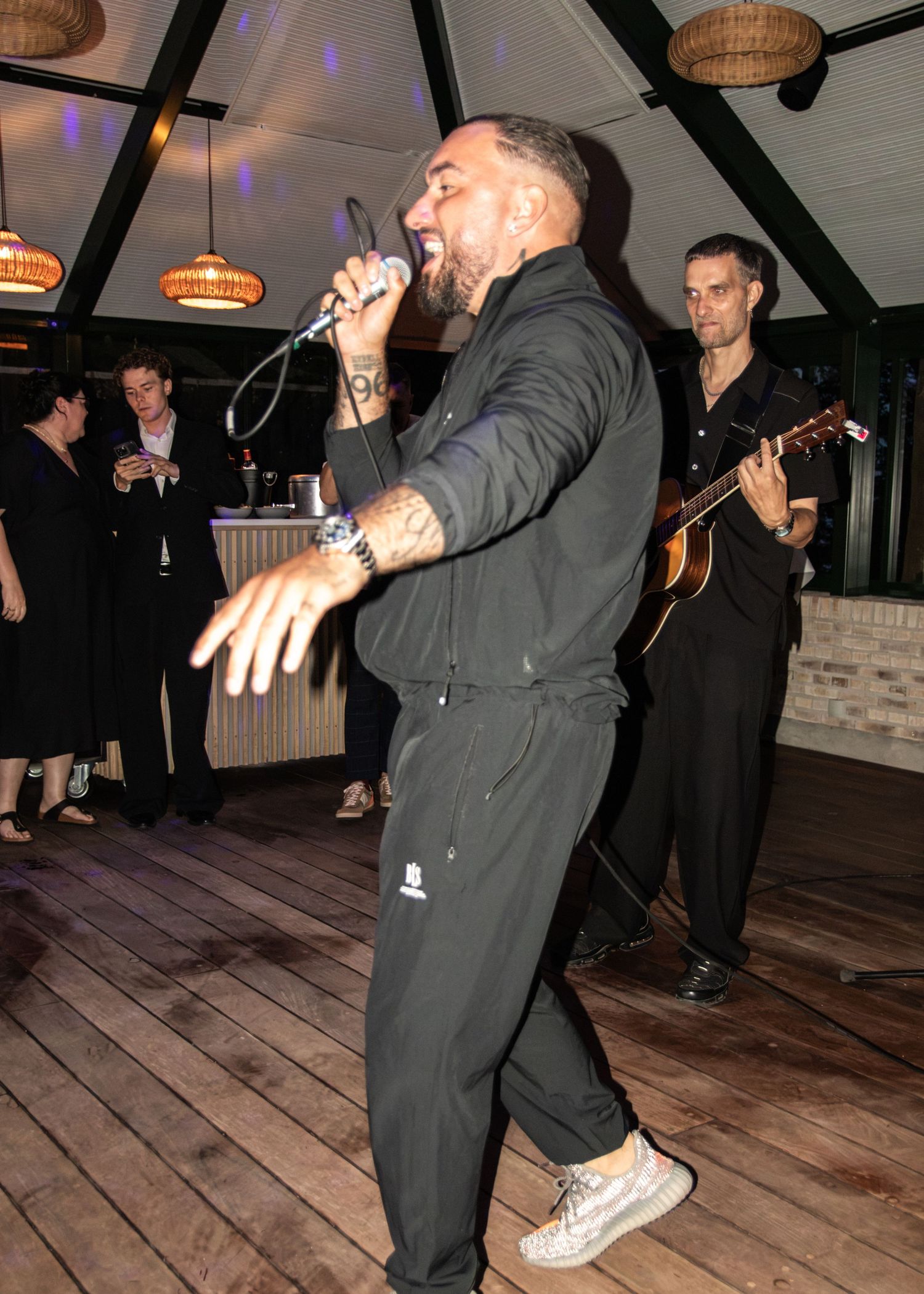 A tattooed singer in a dark outfit performs energetically into a microphone while a guitarist plays at a lively party venue.