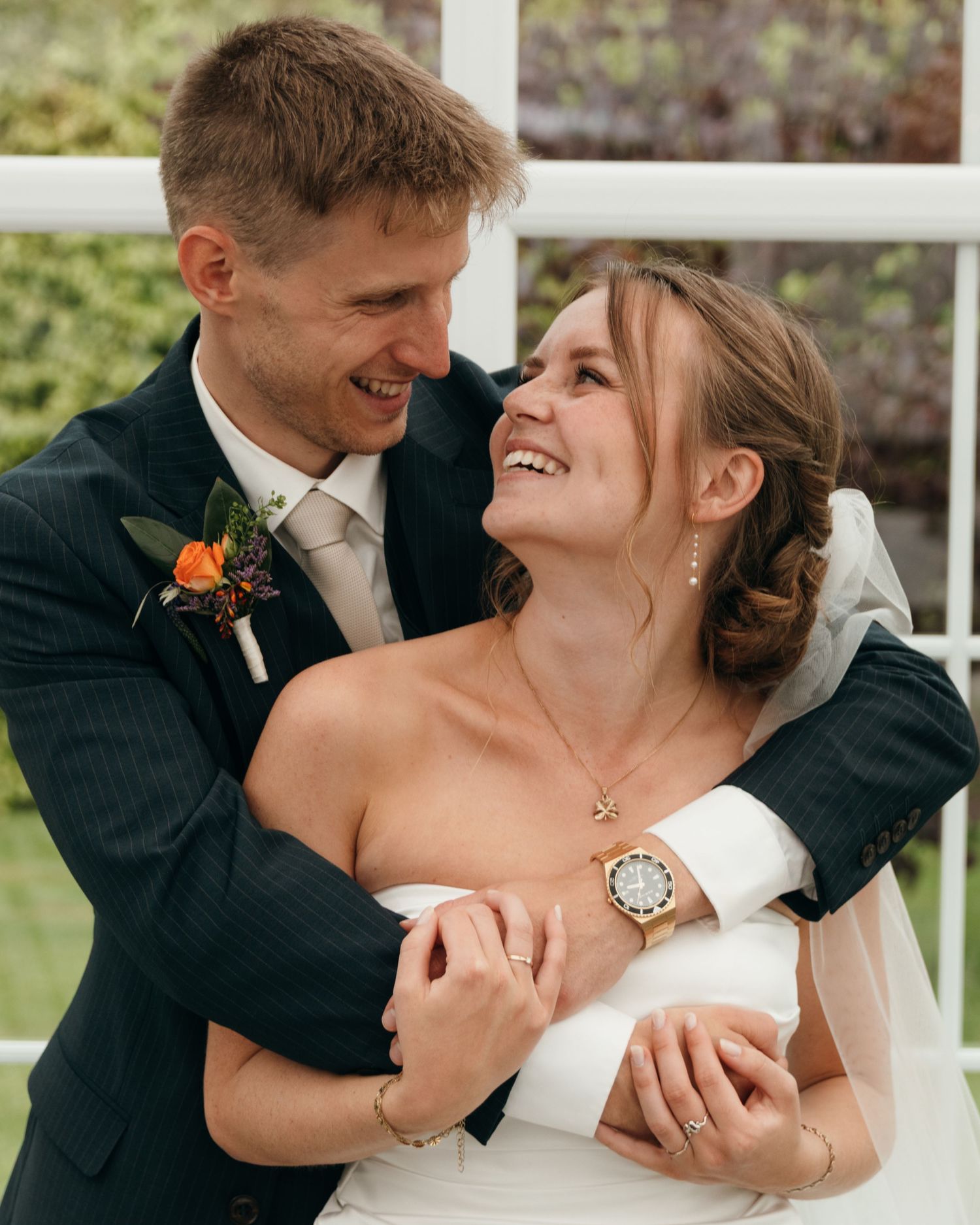 Laughing groom hugs his joyful bride from behind, showing her wristwatch and rings, with an orange boutonniere on his suit.