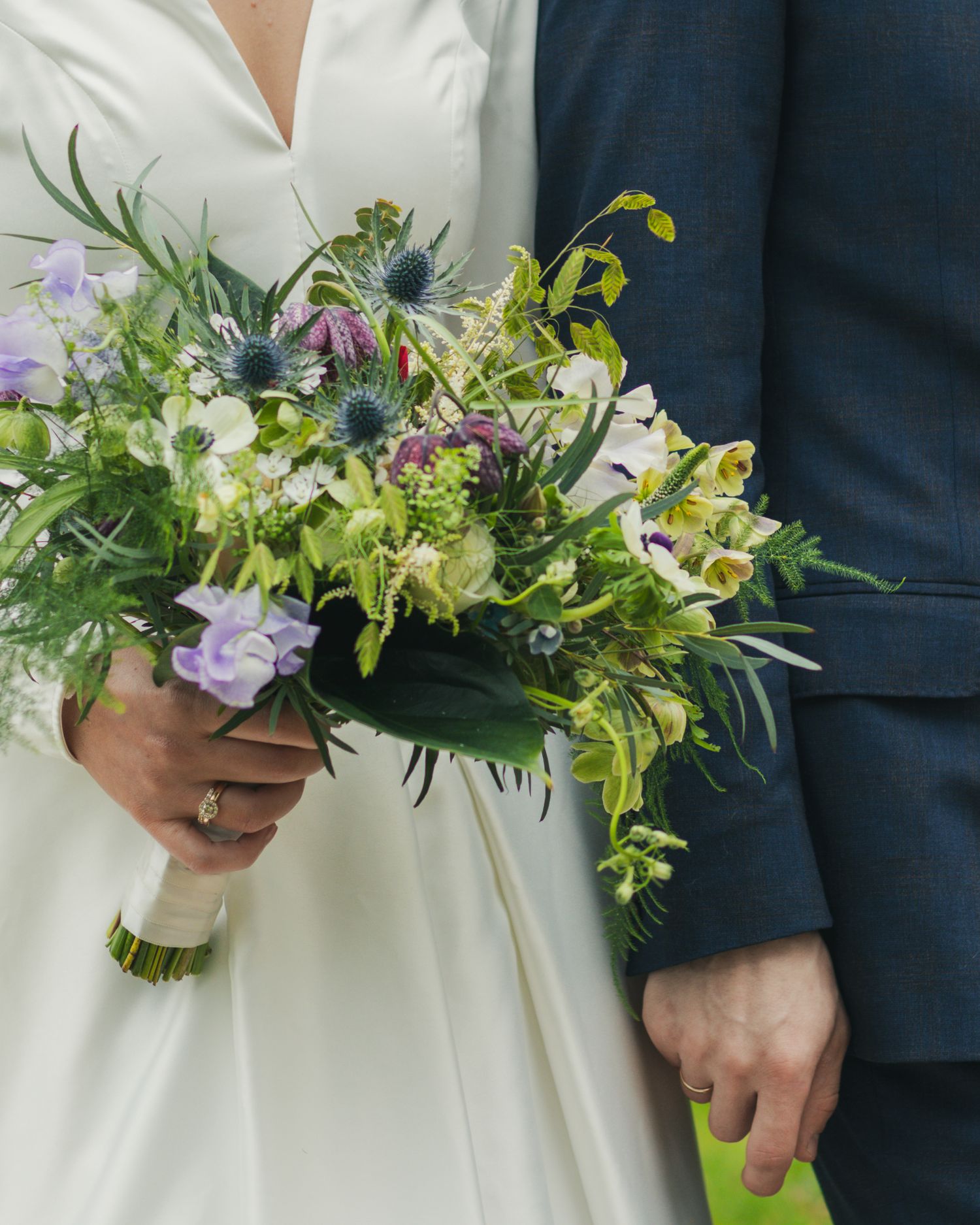 Close-up of a bride holding a wildflower wedding bouquet of thistles, purple blooms and greenery beside her groom in a navy suit.