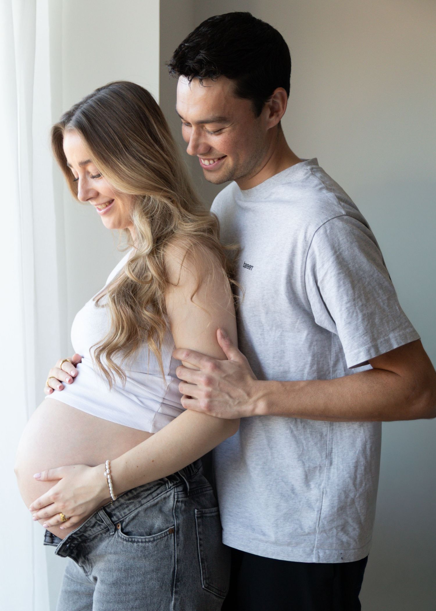 Smiling expectant couple stand by a window as the man gently embraces his pregnant partner, who cradles her baby bump.