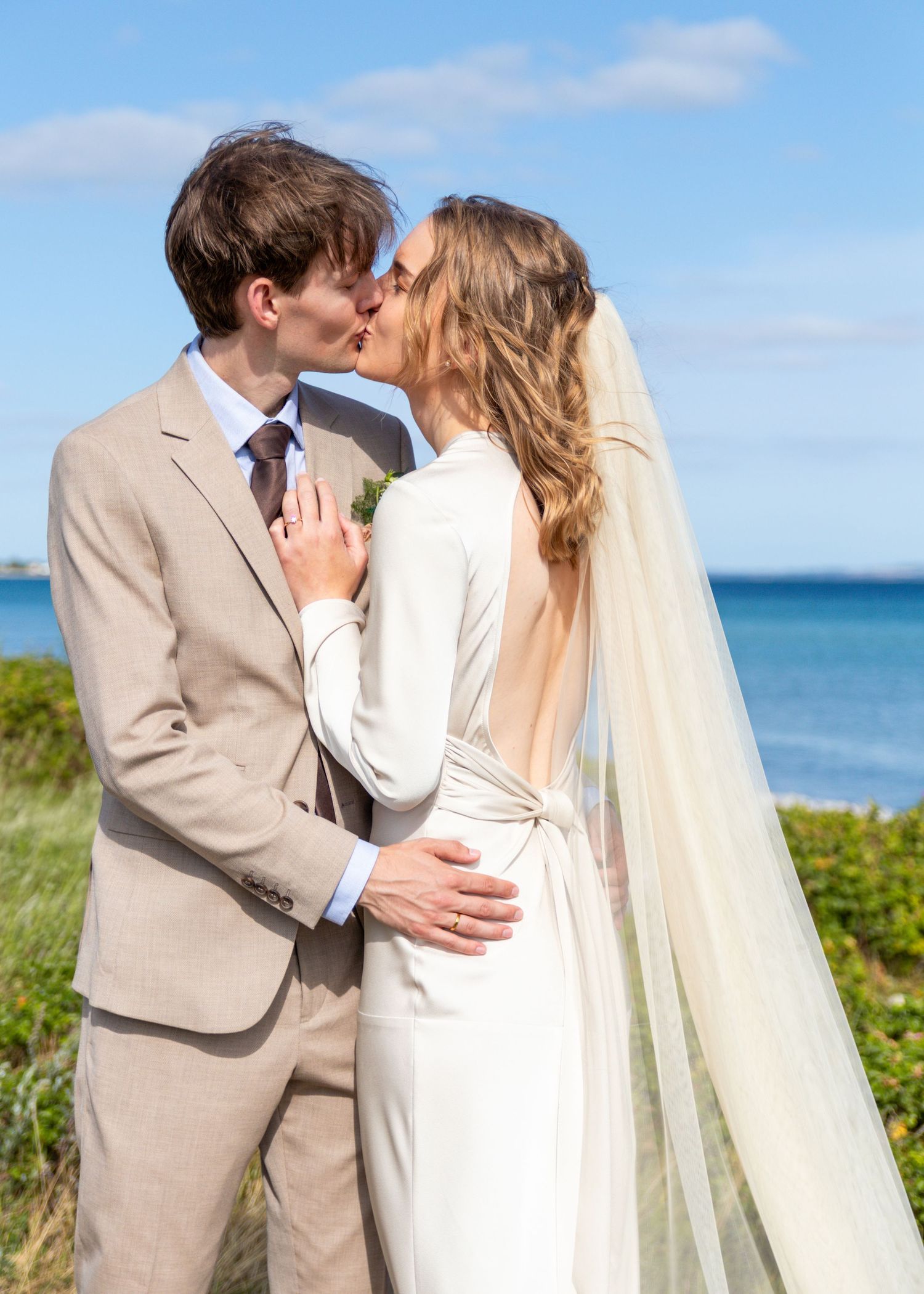 A bride with a long veil and a groom in a beige suit kiss on a grassy coastal bluff overlooking the blue sea.