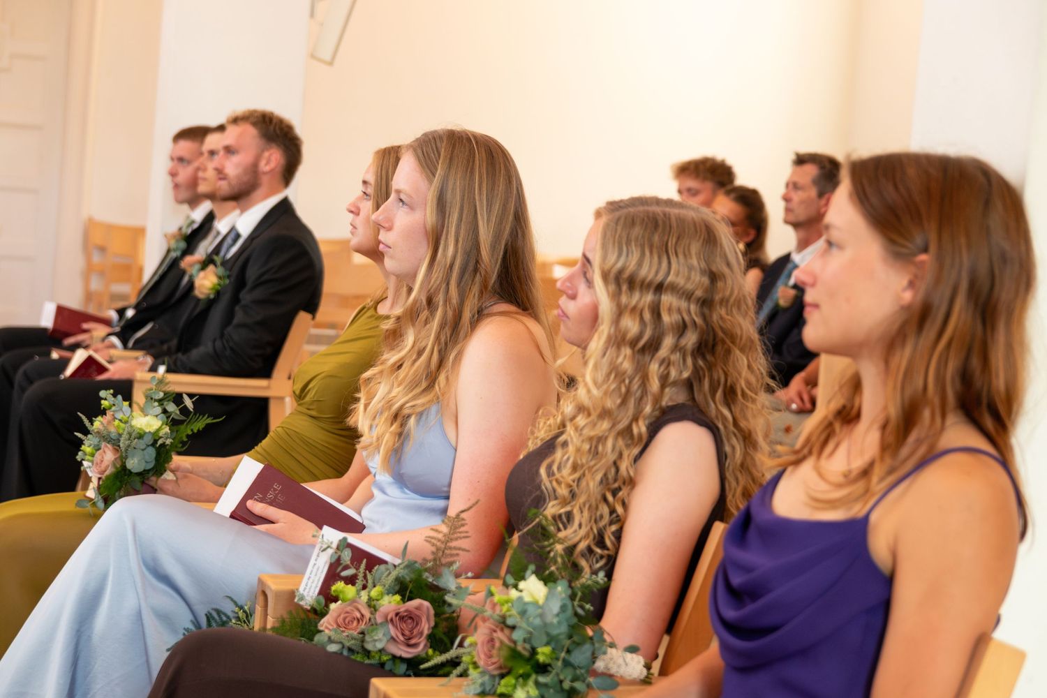 Wedding guests sit in wooden pews holding programs during a ceremony, with flower bouquets resting on the benches.