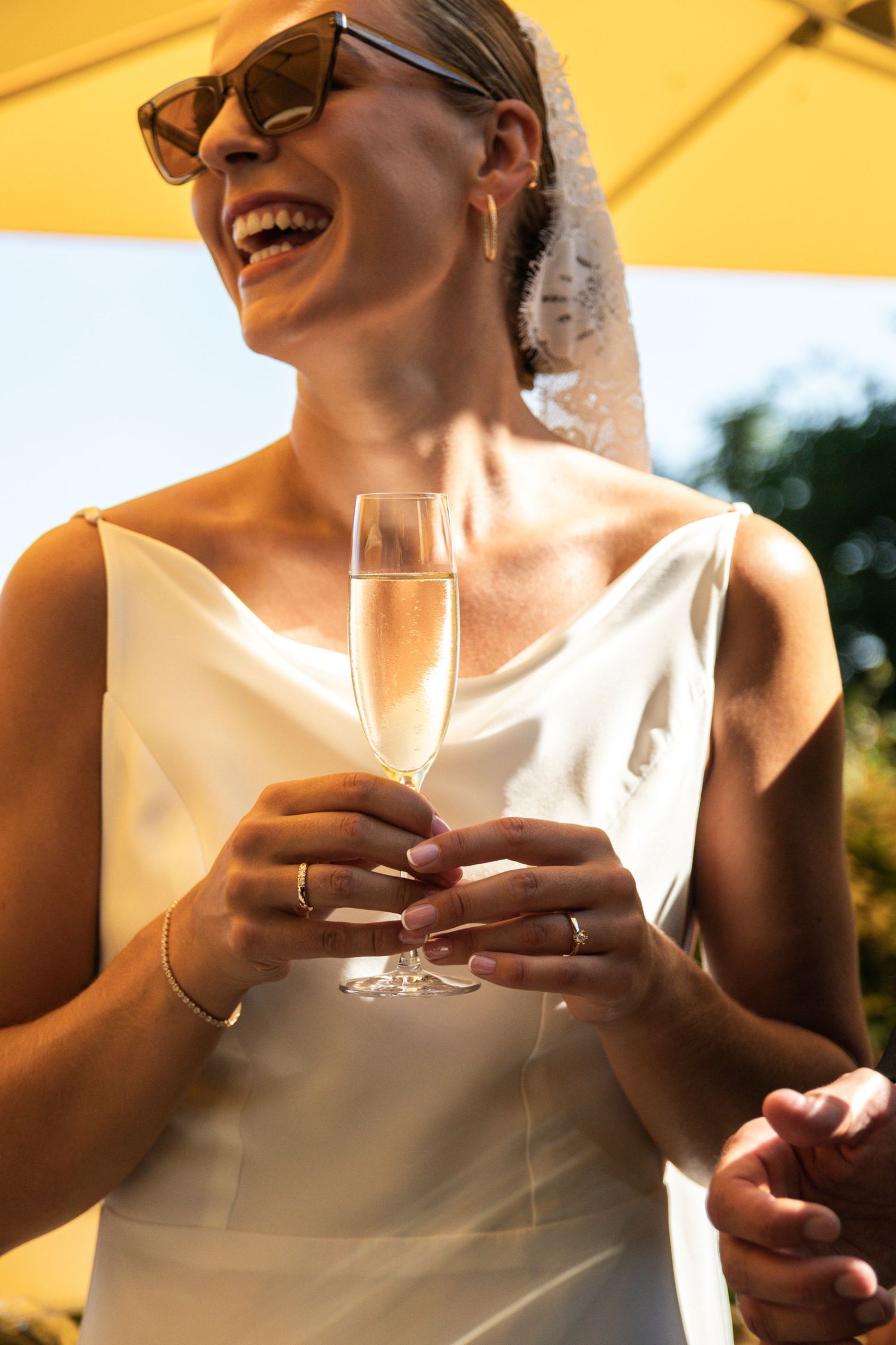 A laughing bride in sunglasses and a lace veil holds a champagne flute during a sunny outdoor wedding reception.
