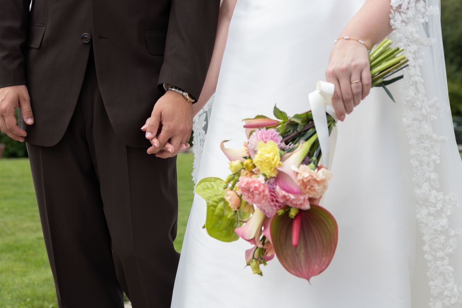 Close-up of a bride and groom holding hands on a lawn, with the bride clutching a bouquet of pink and yellow flowers.