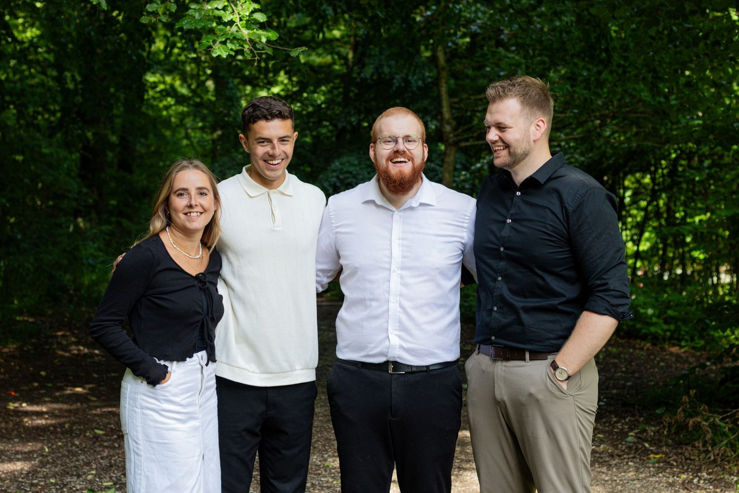 Two men and a woman in smart casual outfits pose together smiling in a green forest for a friendly group portrait.