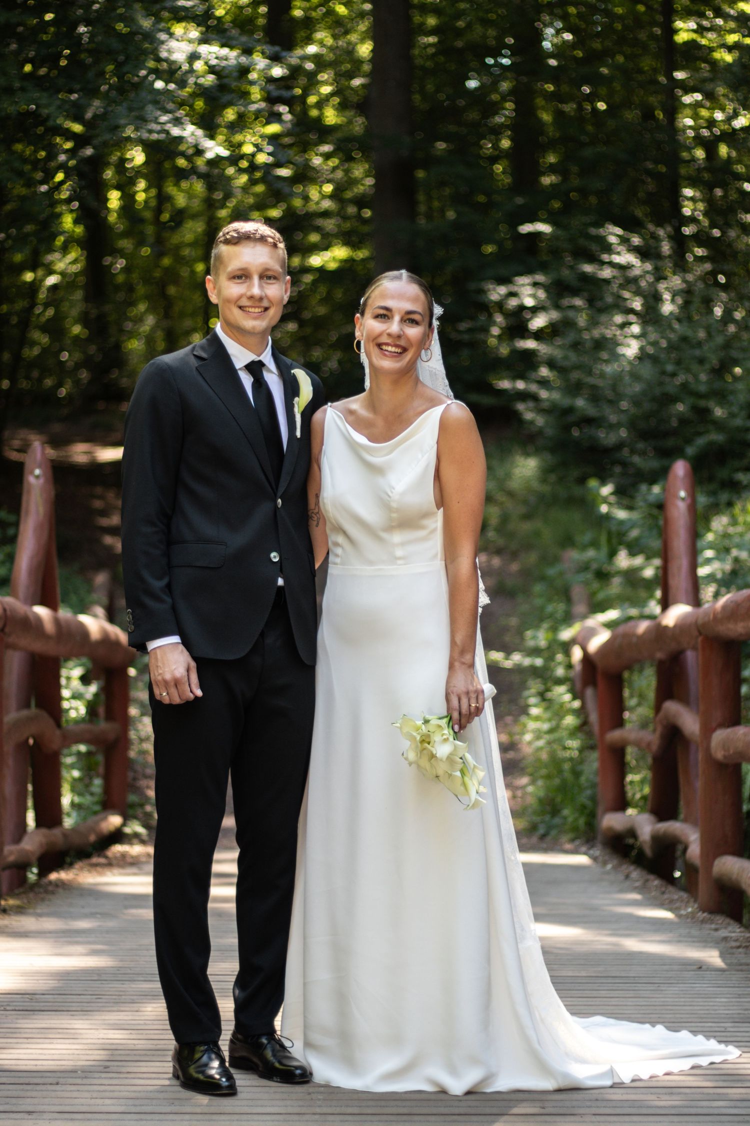 A joyful groom in a black suit stands beside his bride in an elegant white gown and veil on a sunny forest walkway.