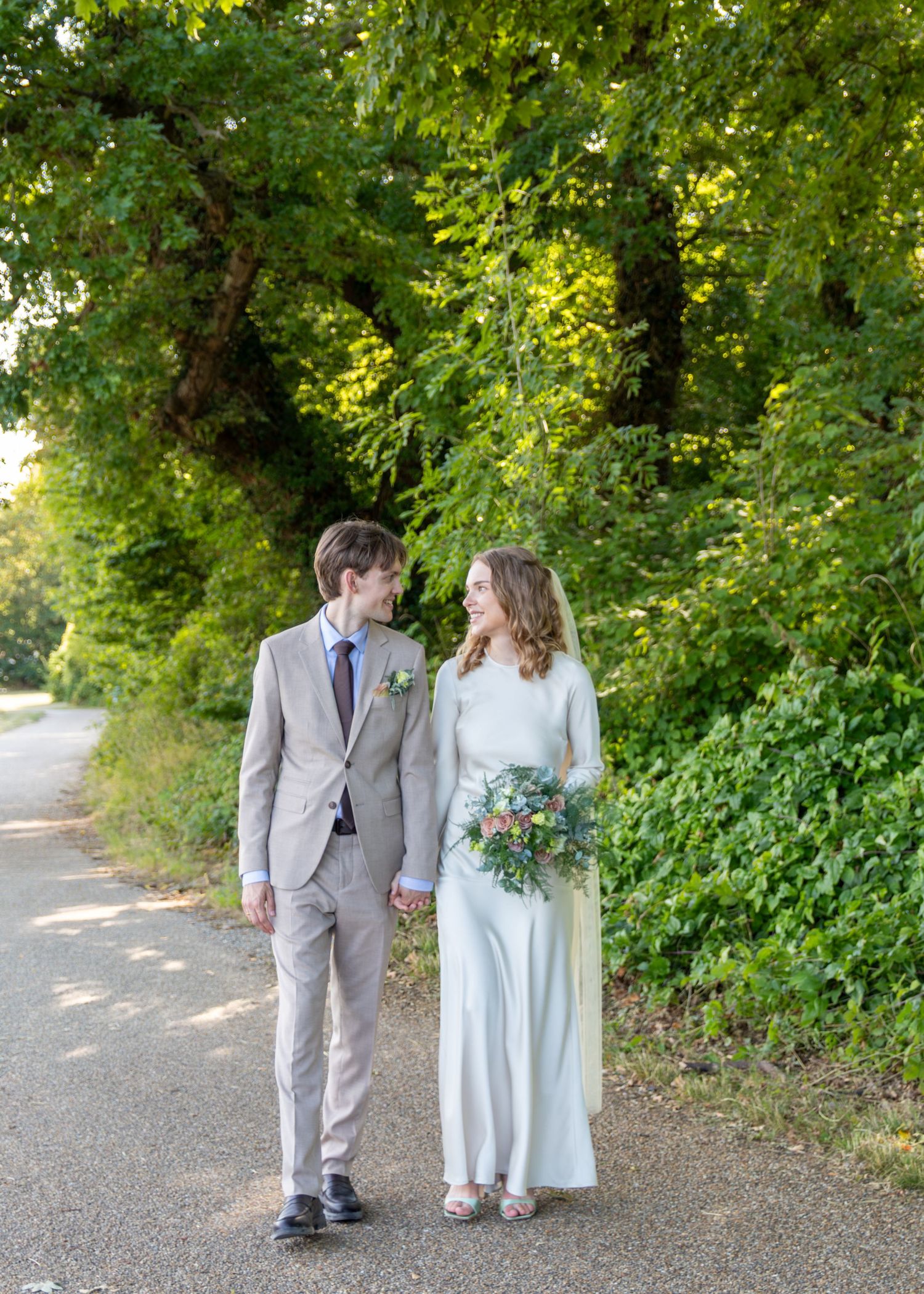 A bride in a satin gown and a groom in a beige suit walk hand in hand along a tree-lined path, smiling at each other.