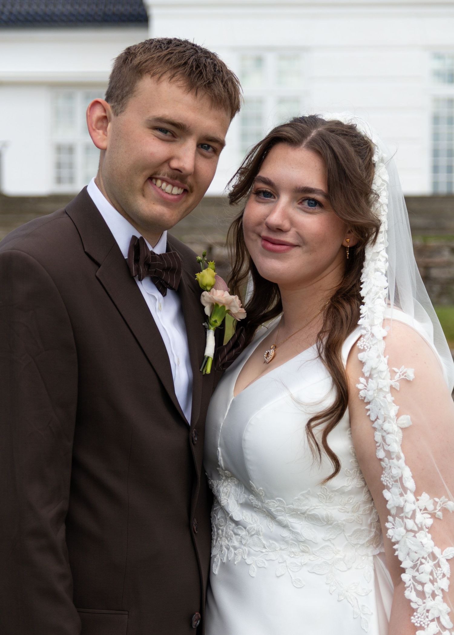 A smiling bride in a lace-trimmed veil and a groom in a brown suit and bow tie pose together outdoors on their wedding day.