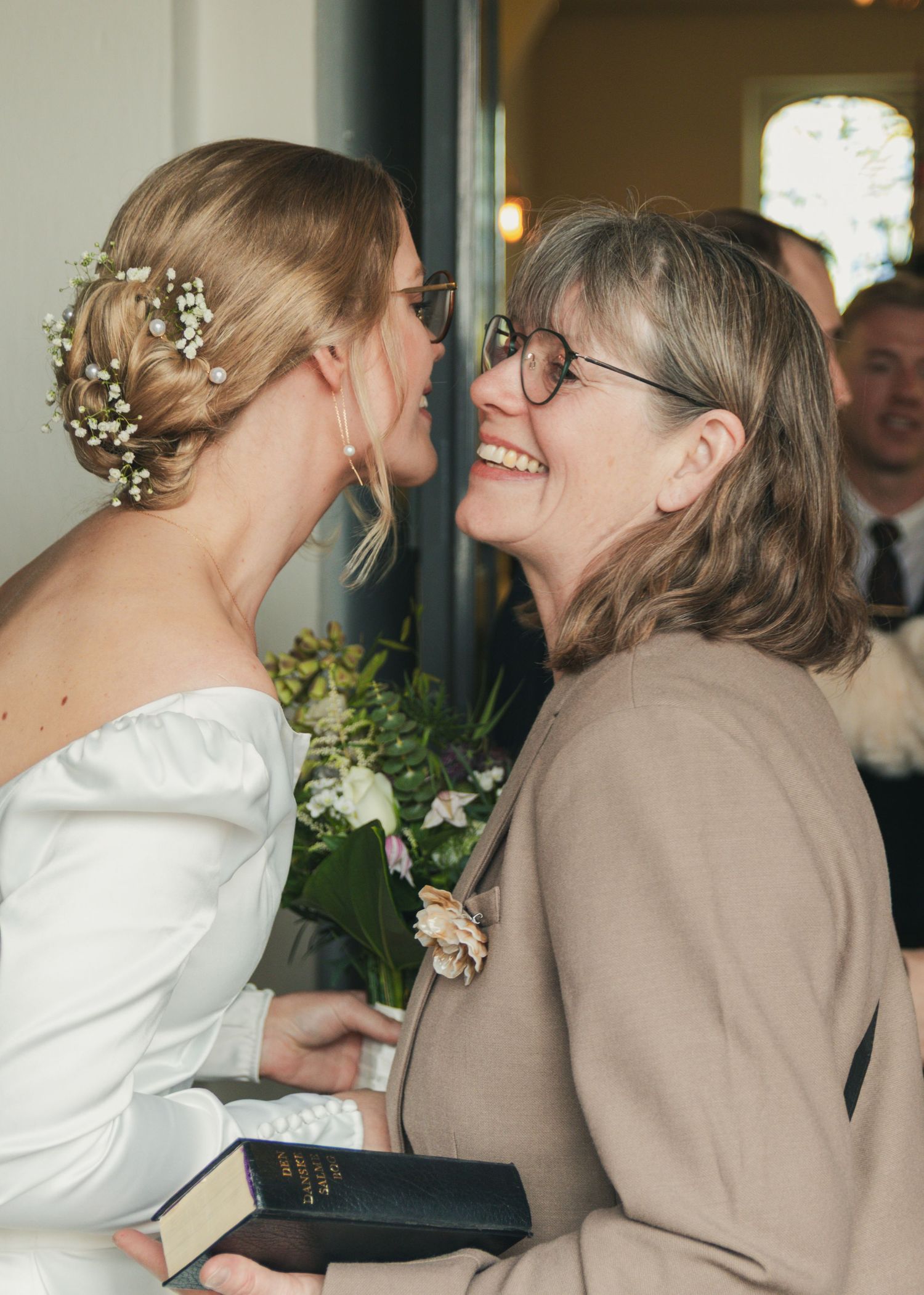 A bride in a white gown with floral hair accessories shares a joyful moment with a smiling woman in glasses holding a black book.