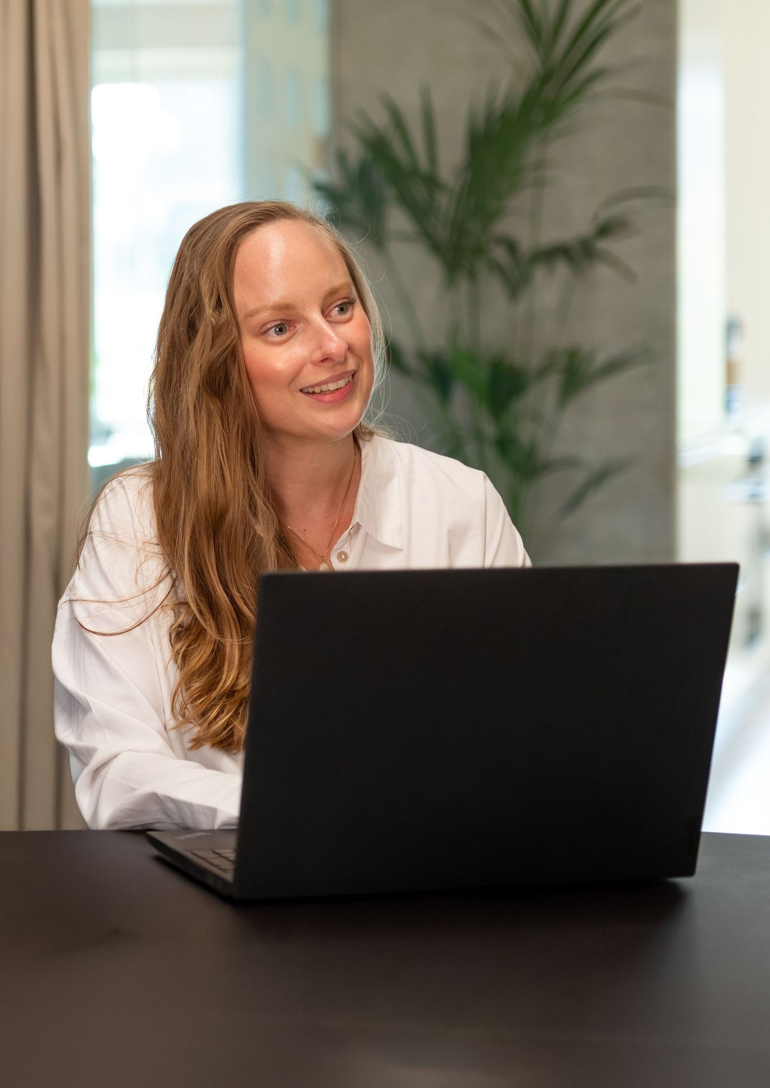 A smiling woman with long hair in a white blouse sits at a dark table with a laptop beside a leafy green plant.