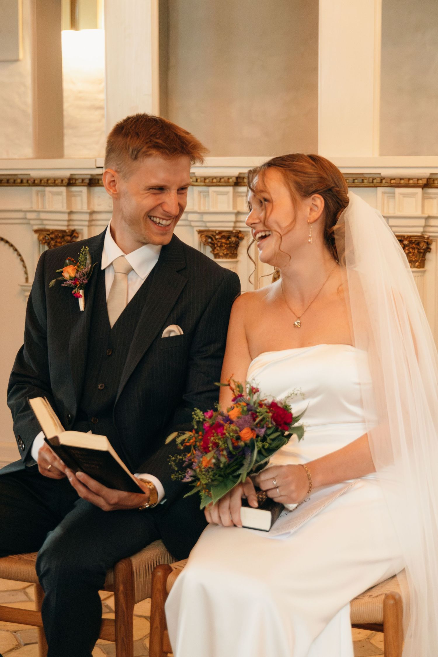 Bride and groom laugh joyfully while seated in a church, holding a book and a bright floral bouquet on their wedding day.