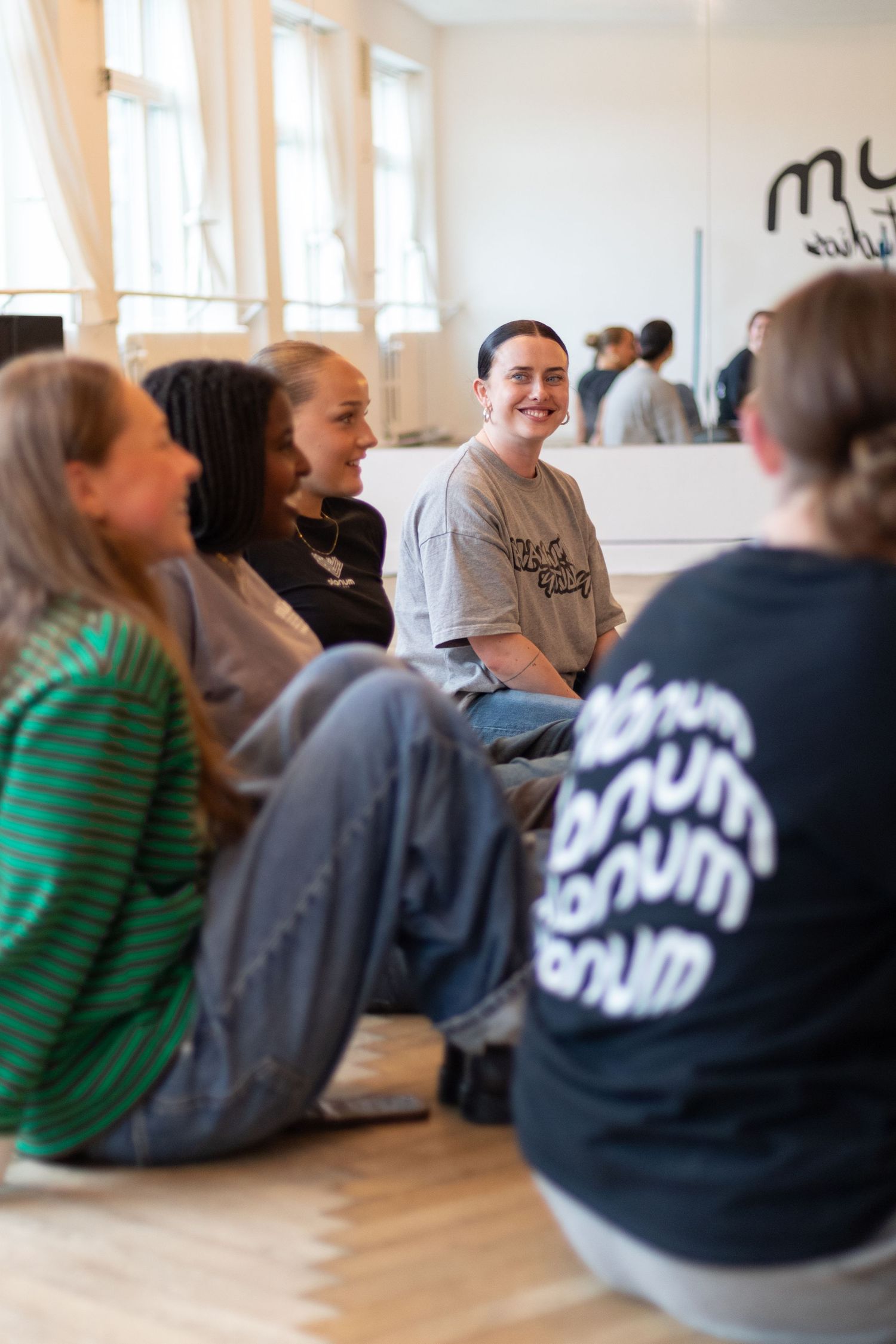 Group of dancers sits on a wooden floor in a bright studio, smiling and listening during a dance class.