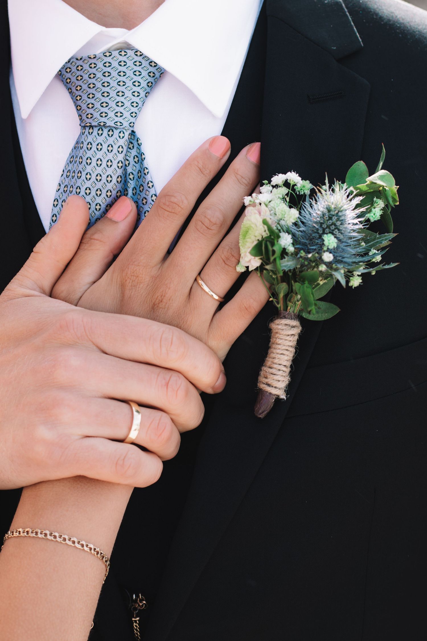 Close-up of the couple's hands with wedding rings resting on the groom's suit beside a blue thistle boutonniere and patterned tie.