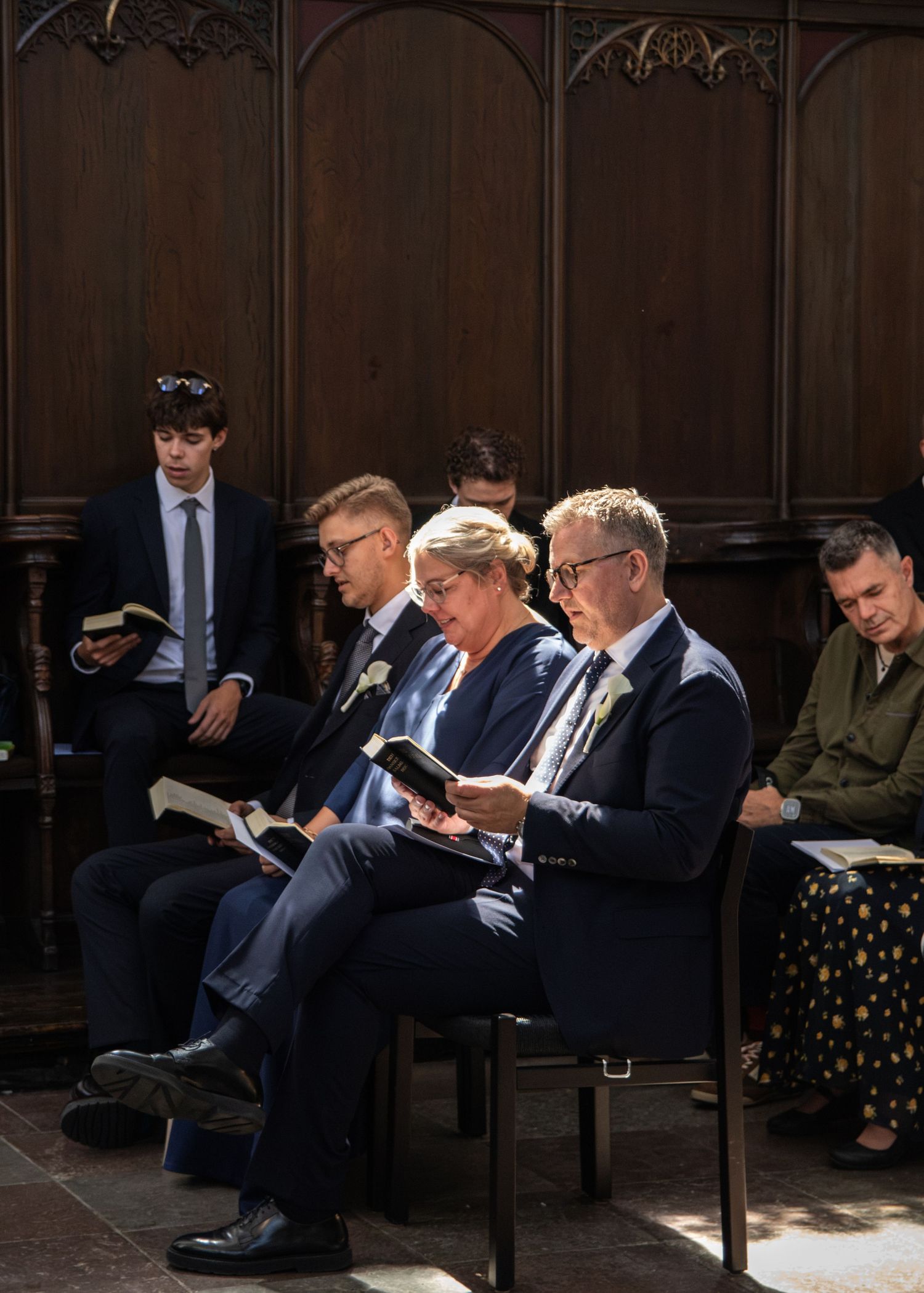 Wedding guests in formal attire sit in carved wooden pews reading hymn books during a church service.
