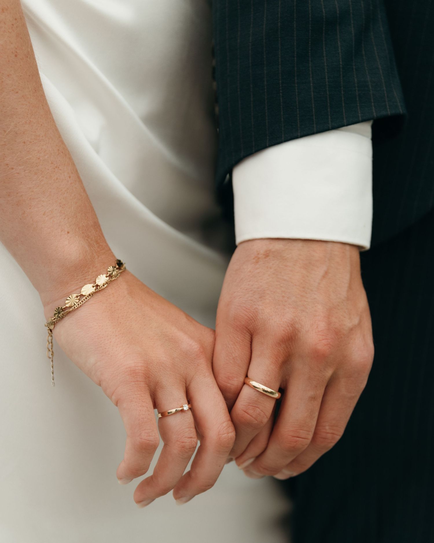 Close-up of a bride and groom holding hands, showing gold wedding rings, a delicate bracelet, and his pinstripe suit sleeve.