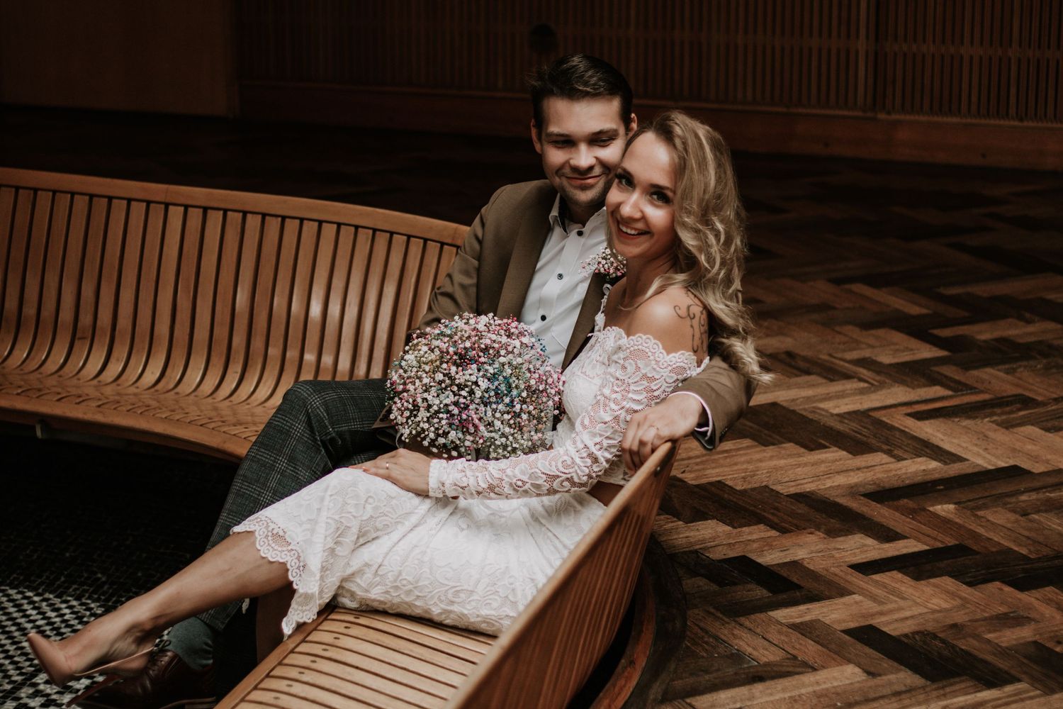 A smiling bride in a white lace dress holds a colorful bouquet while sitting on a curved wooden bench beside her groom.